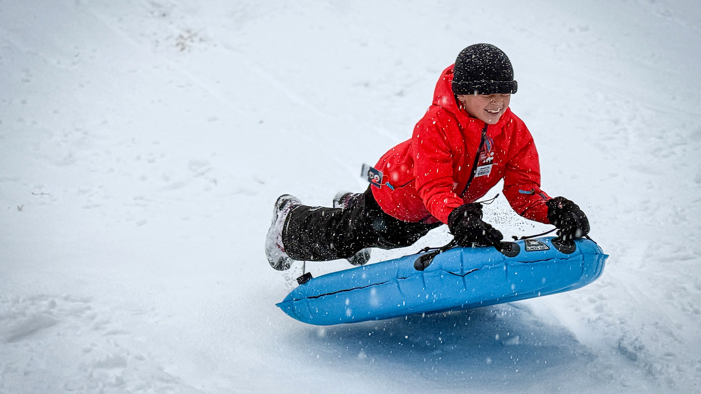 A child in a bright red winter jacket laughs while sledding down a snowy hill, airborne above a raft-type sled, photographed for a story by author Jakob Schiller.