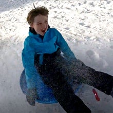 A child in a bright blue winter jacket laughs while sledding down a snowy hill, boots kicked forward as snow sprays up around them, photographed for a story by author Jakob Schiller.