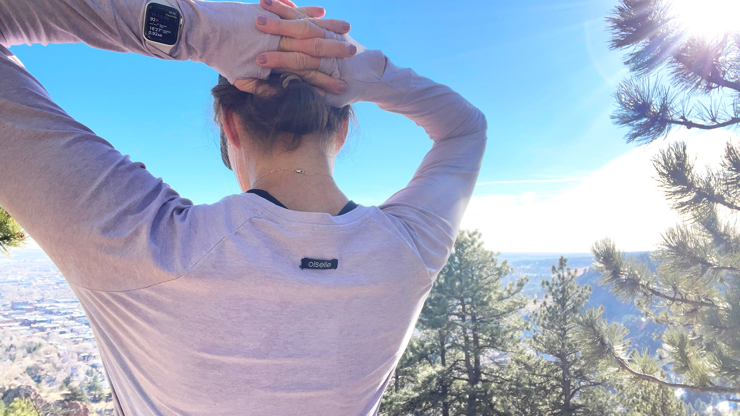 A runner photographed by Lisa Jhung wears a lavender long-sleeve Oiselle shirt, showcasing how to use breathable running layers to maintain temperature during an outdoor workout. The image shows the back of the runner against a clear blue sky, highlighting the shirt's thumbholes and lightweight fabric.