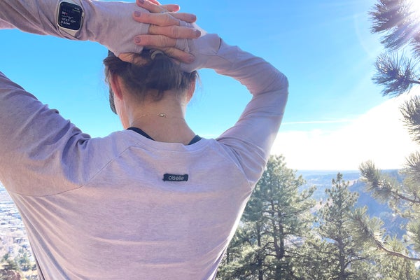A runner photographed by Lisa Jhung wears a lavender long-sleeve Oiselle shirt, showcasing how to use breathable running layers to maintain temperature during an outdoor workout. The image shows the back of the runner against a clear blue sky, highlighting the shirt's thumbholes and lightweight fabric.