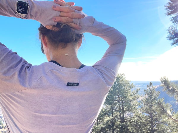 A runner photographed by Lisa Jhung wears a lavender long-sleeve Oiselle shirt, showcasing how to use breathable running layers to maintain temperature during an outdoor workout. The image shows the back of the runner against a clear blue sky, highlighting the shirt's thumbholes and lightweight fabric.