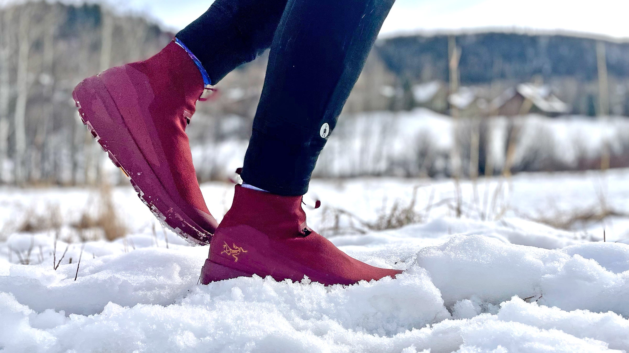 A runner steps through fresh snow wearing burgundy Arc’Teryx Nivalis winter running shoes with grippy soles and insulated uppers, paired with black tights, in a cold, wintry landscape; photo by Lisa Jhung.