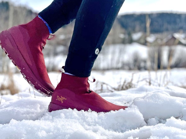 A runner steps through fresh snow wearing burgundy Arc’Teryx Nivalis winter running shoes with grippy soles and insulated uppers, paired with black tights, in a cold, wintry landscape; photo by Lisa Jhung.