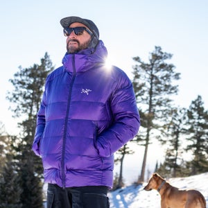 Outside Contributor Jakob Schiller stands outdoors in a snowy mountain landscape wearing a bright purple Arc'teryx jacket, black pants, sunglasses, and a cap, with evergreen trees and a dog in the background as sunlight flares behind him.