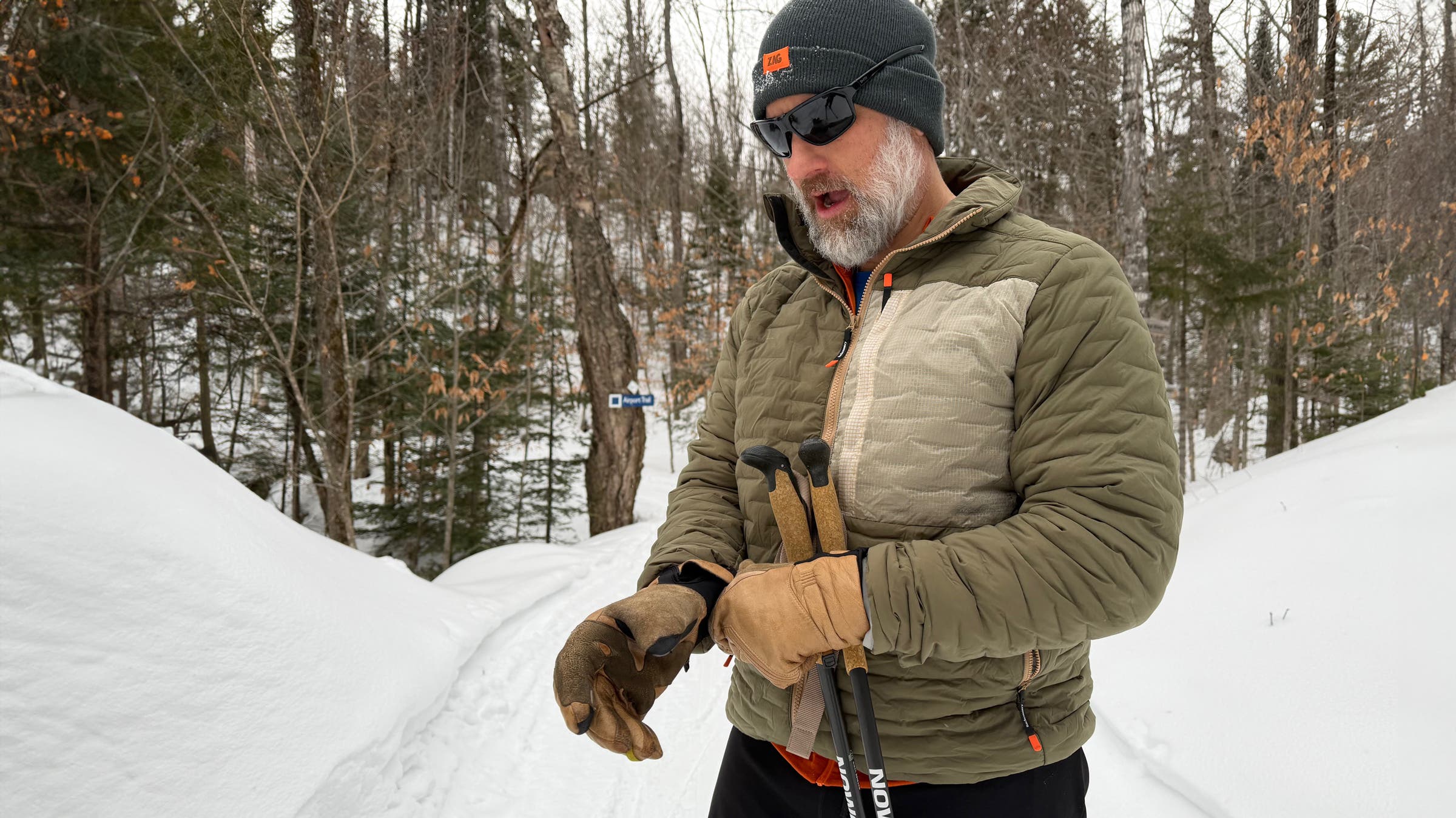 A man adjusts his gloves on a snowy forest trail while wearing the Backcountry Peale Primaloft Stitch-Free Baffle Stretch Jacket, a lightweight synthetic jacket designed for cold-weather hiking and winter layering.