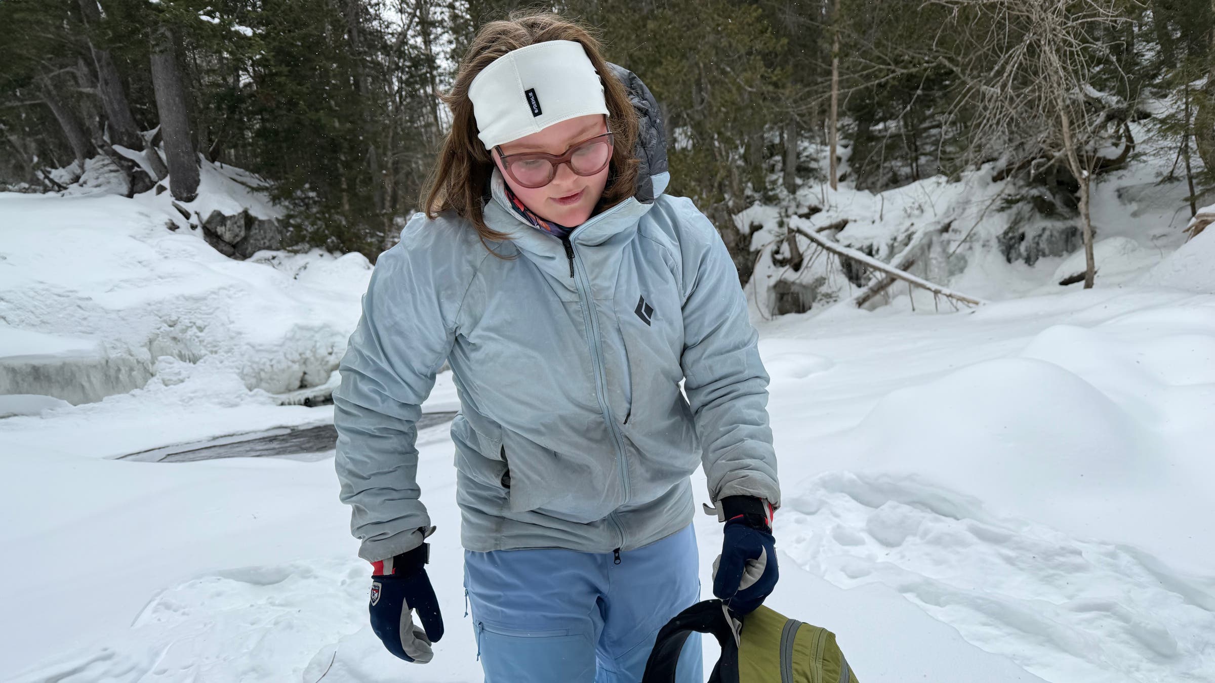 A hiker navigates a snowy creekside trail while wearing the Black Diamond Solution 4.0 Parka, a durable synthetic jacket built to provide warmth and weather protection during cold winter adventures.