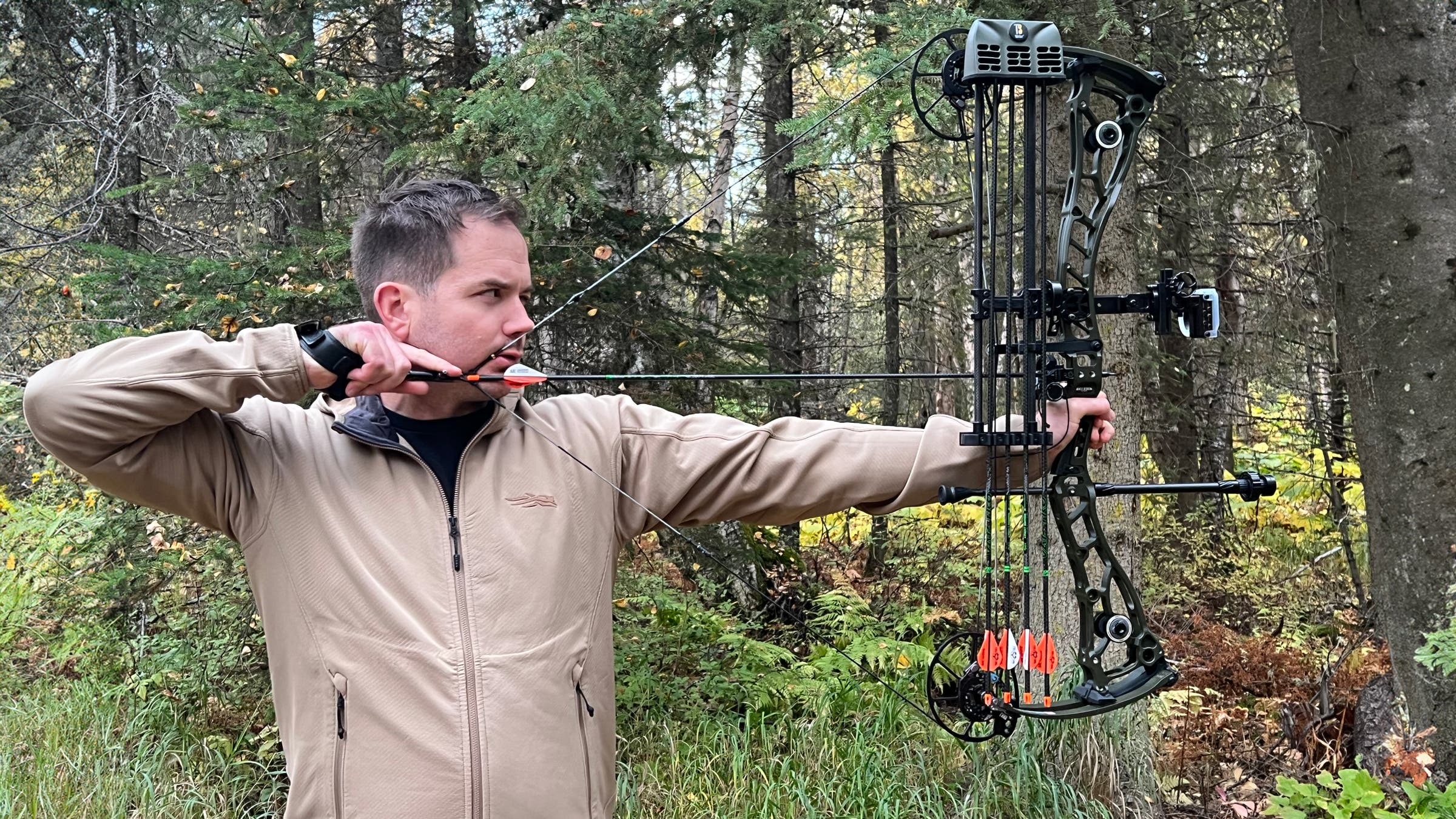 Man drawing a Bowtech Proven 34 compound bow at full draw in a wooded forest setting, aiming an arrow with mounted sight and stabilizer, showcasing premium hunting accessories in action.