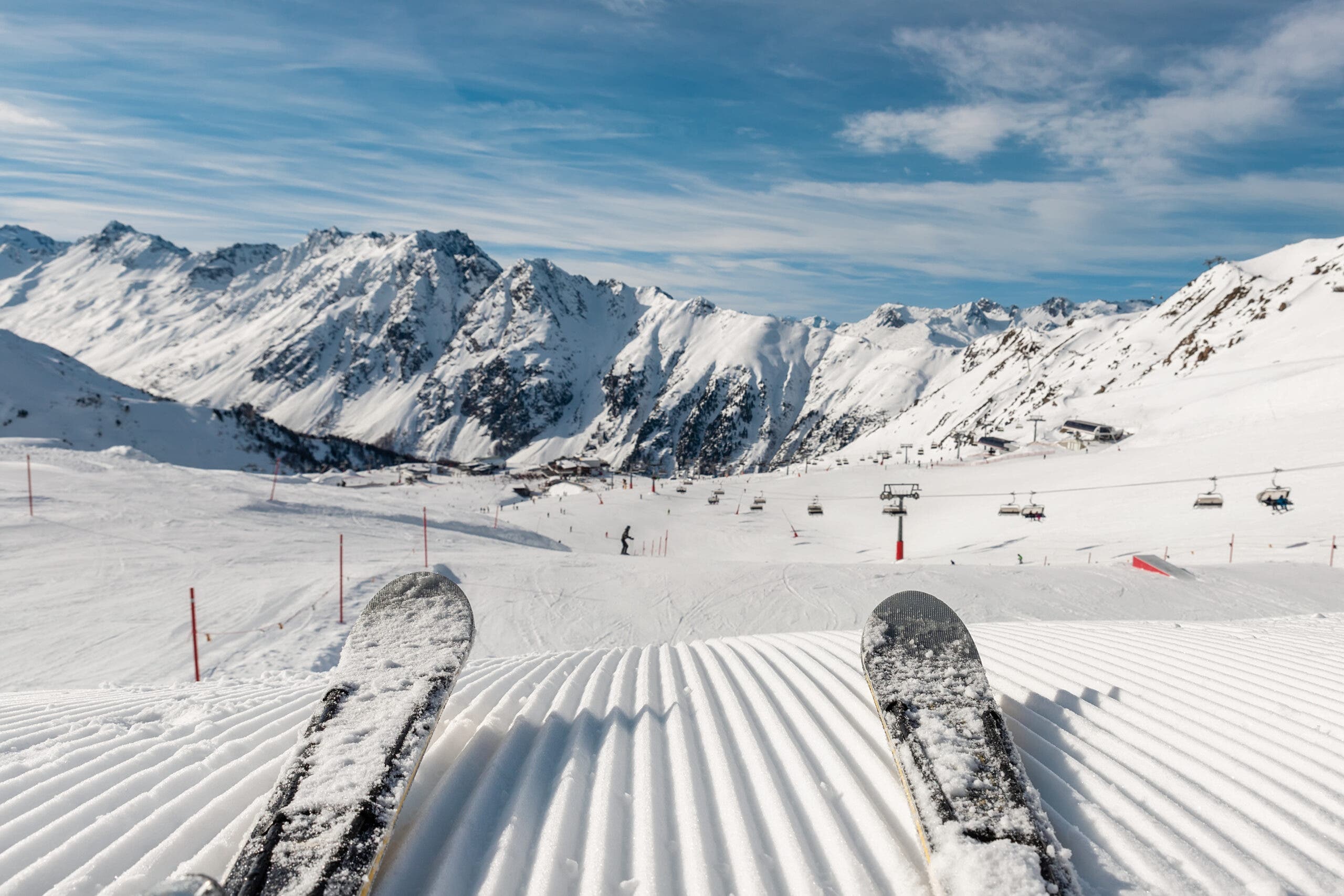 Panoramic point of view skier legs on downhill start straight line rows freshly prepared groomed ski slope piste on bright day blue sky background. Snowcapped mountain landscape europe winter resort.