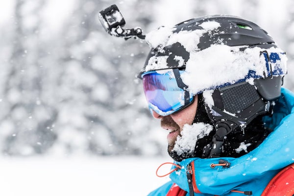 Closeup headshot of skier wearing black GoPro helmet camera in front of snowy background