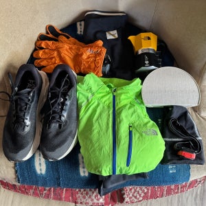 Flat lay of gear to use in the dark for workouts, including black running shoes, a bright green running jacket, orange insulated gloves, a headlamp, reflective accessories, and small essentials arranged on a patterned blanket indoors.