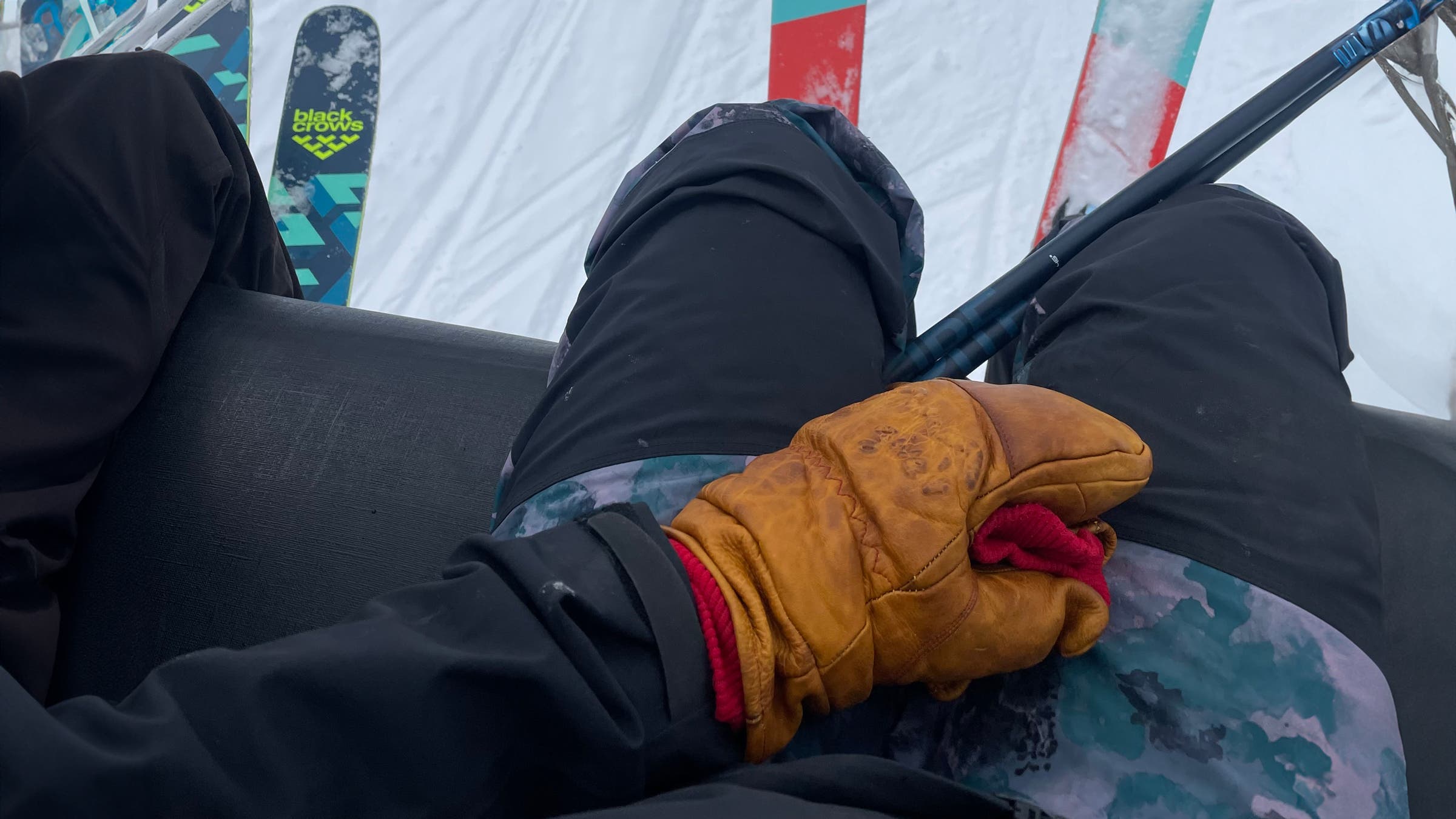 Close-up of a skier riding a chairlift wearing a well-broken-in Give’r Frontier Mitten with red knit cuff, resting on their lap beside ski poles and skis in the background.