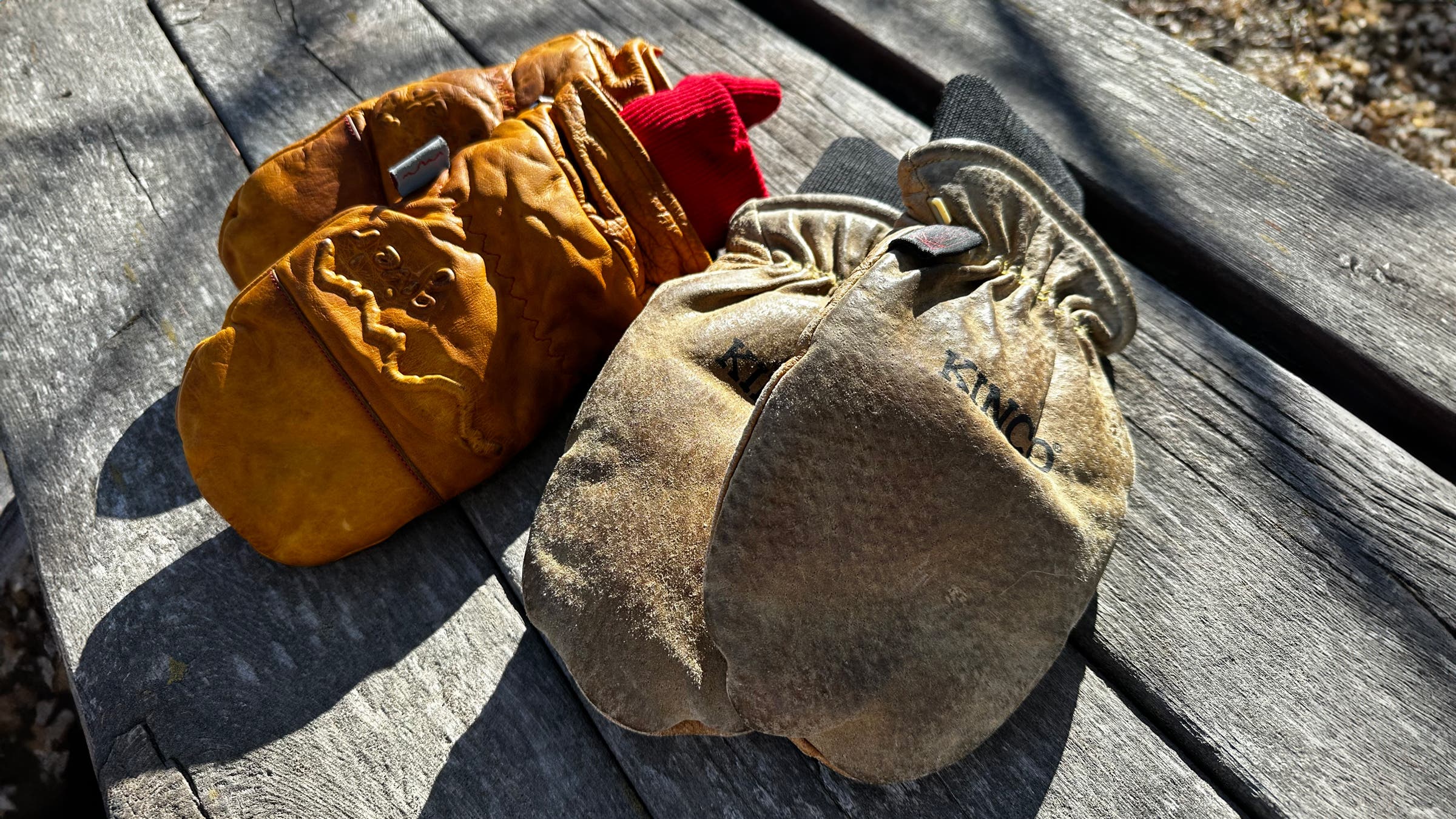 Two pairs of leather mittens—the golden-brown Give’r Frontier with red knit cuffs and the weathered tan Kinco Grain & Suede with black cuffs—rest side by side on a sunlit wooden picnic table, highlighting their broken-in texture and rugged patina