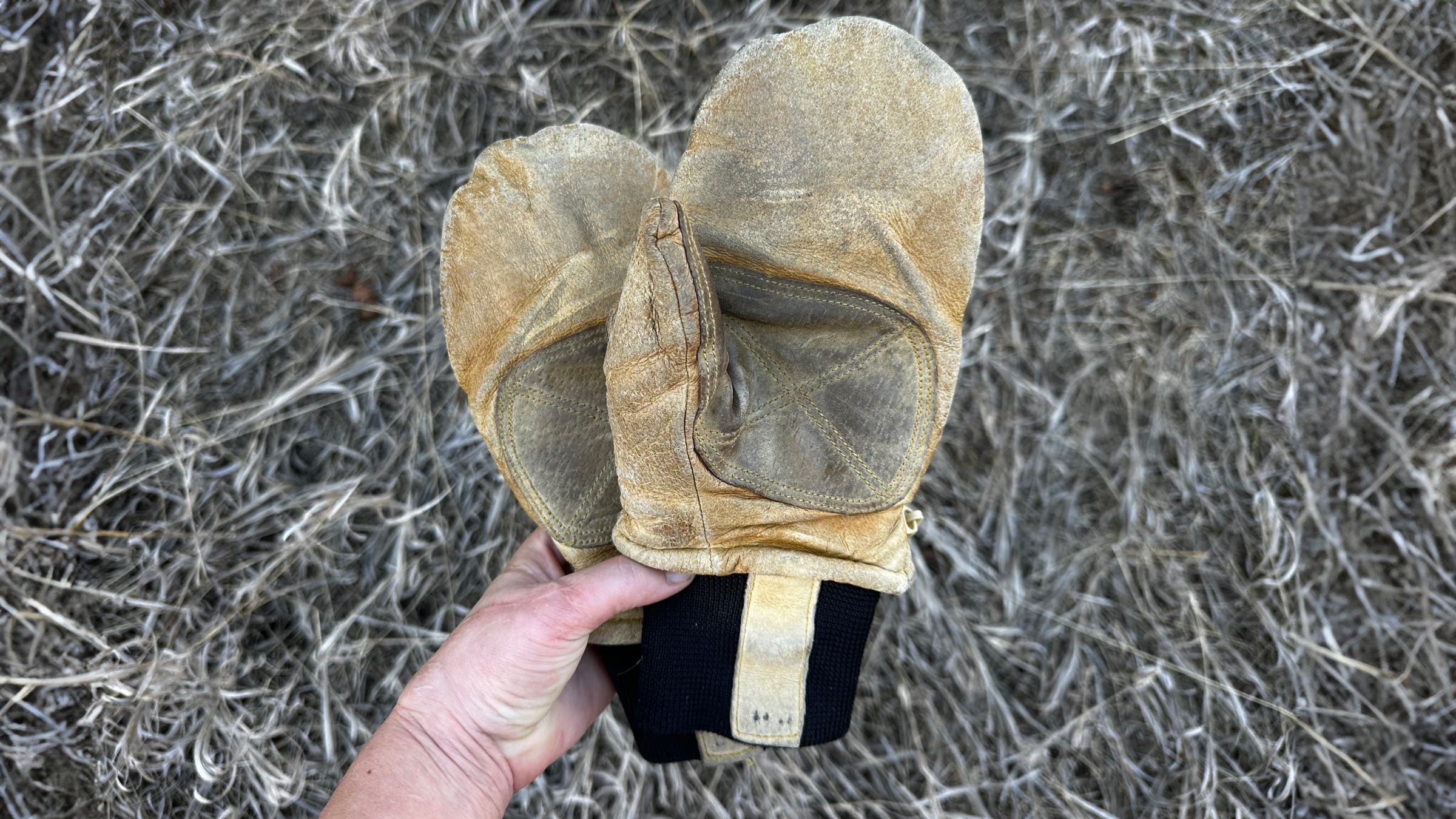 Hand holding a well-worn pair of Kinco Grain & Suede leather mittens over dry grass, showing the reinforced suede palm patches, creased grain leather, and black knit cuffs.