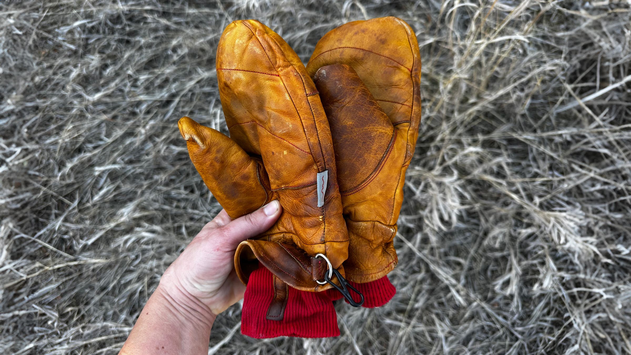 Hand holding a well-worn pair of Give’r Frontier Mitten leather mitts with rich brown patina, visible creases, red knit cuffs, and attached wrist leash, set against dry grass.