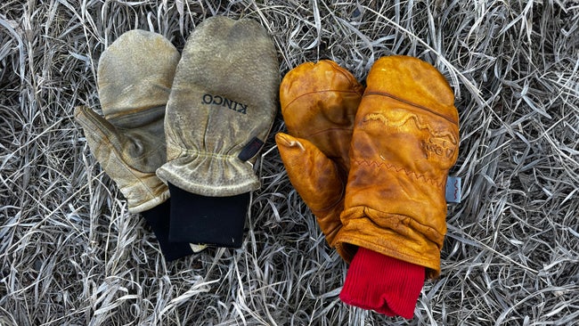 Close-up of two pairs of well-worn leather mittens resting on dry grass: the tan Give’r Frontier mitts with red knit cuffs on the right and the lighter Kinco Grain & Suede mitts with black cuffs on the left, showing natural creases and patina from heavy winter use.