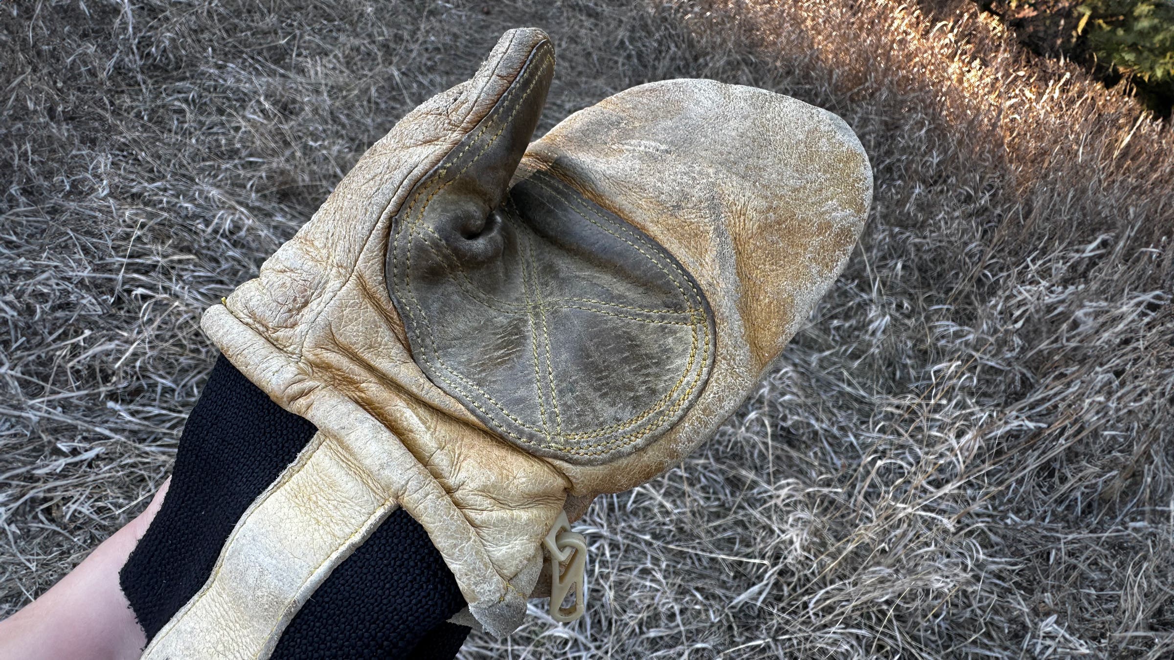 Close-up of a hand holding a Kinco Grain & Suede leather mitten, highlighting the reinforced suede palm patch, heavy stitching, creased grain leather, and snug black knit cuff against a backdrop of dry grass.
