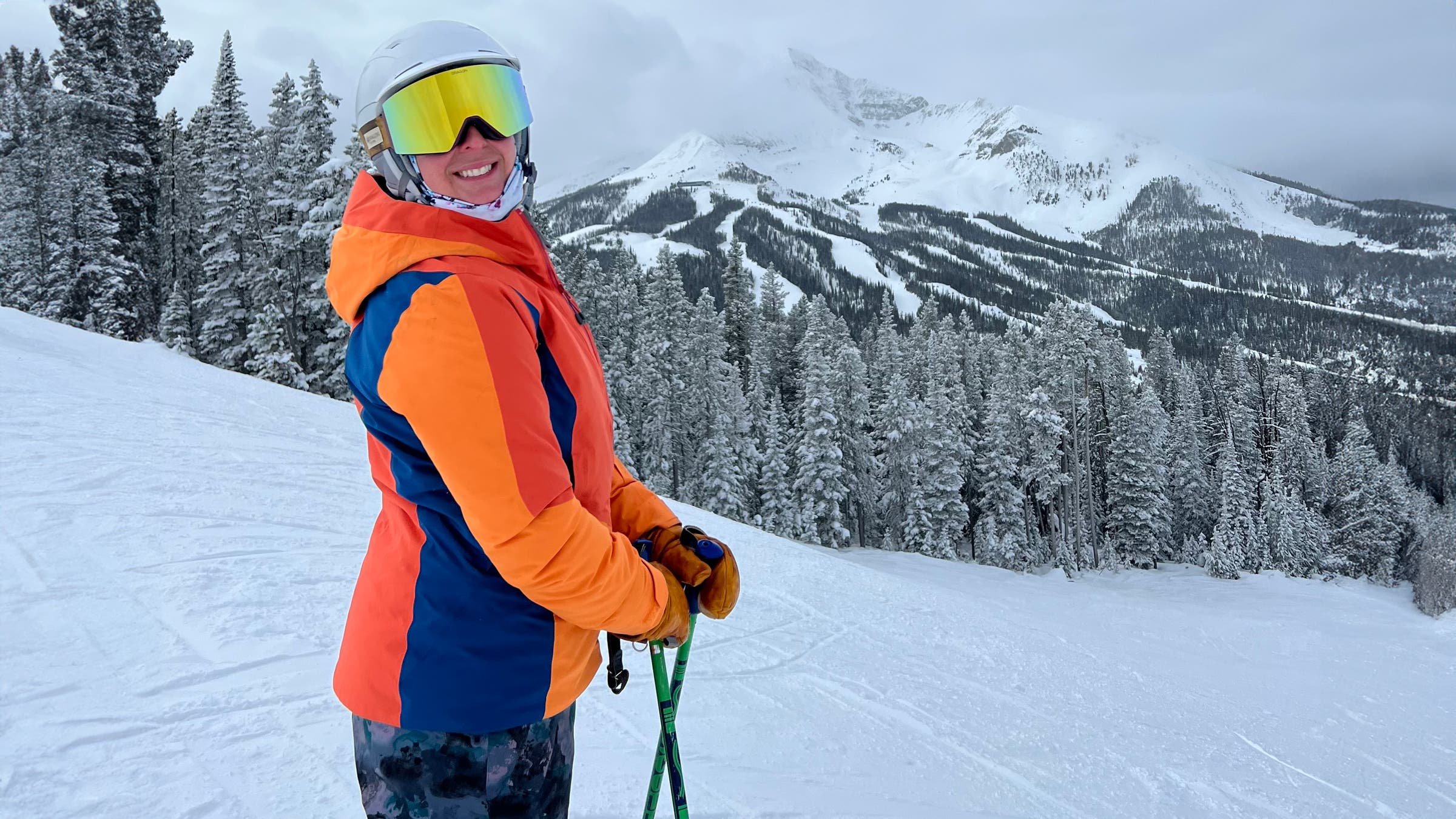 Smiling skier in a bright orange and blue jacket stands on a snowy mountain slope wearing Give’r Frontier Mitten leather mitts, holding ski poles with snow-covered trees and rugged alpine peaks in the background.