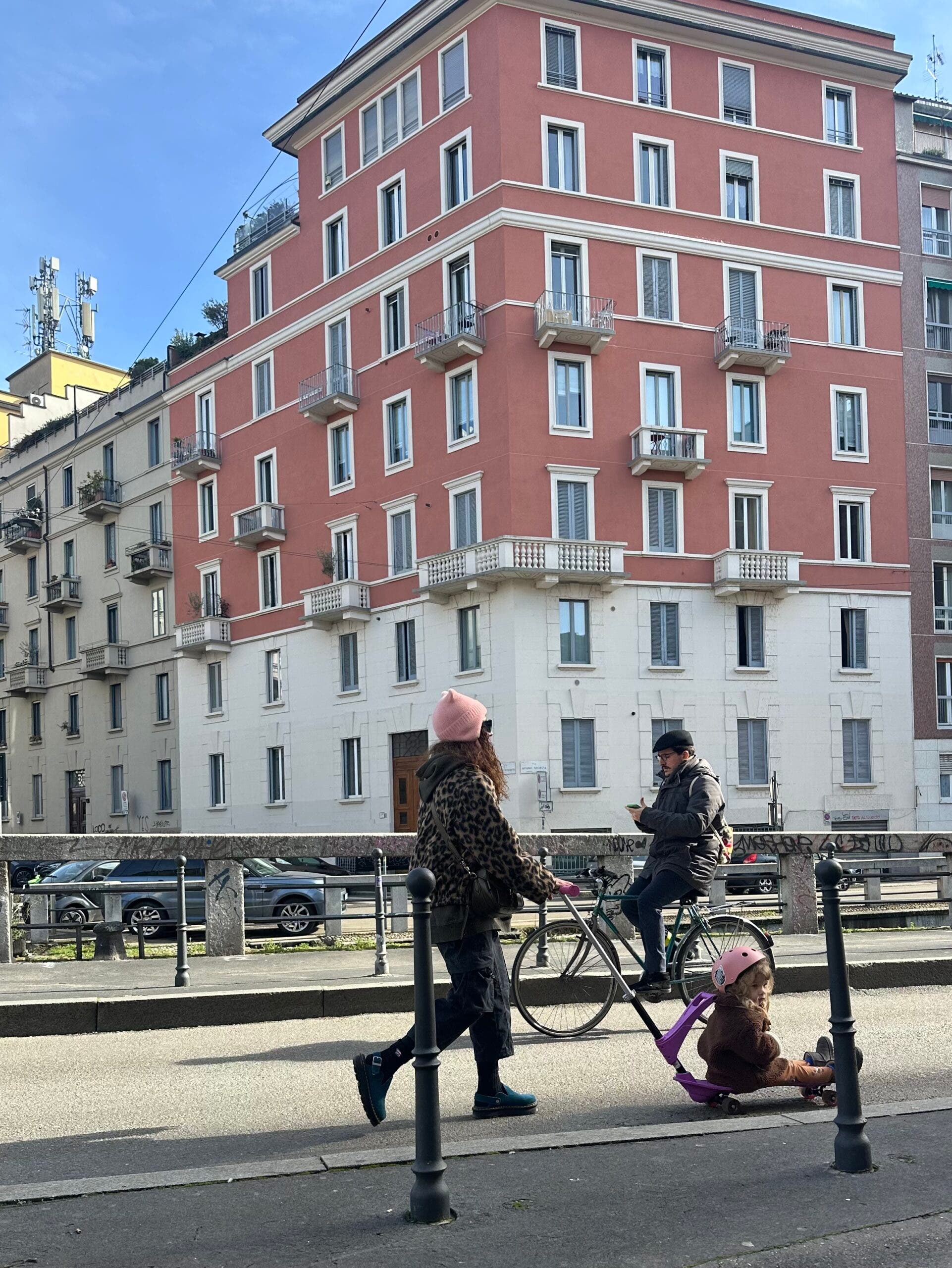 A person walks past a terracotta building in Milan wearing a leopard print fleece, a pink beanie, and dark cargo pants. They are pulling a small child on a purple scooter during the Milan-Cortina Winter Olympics.