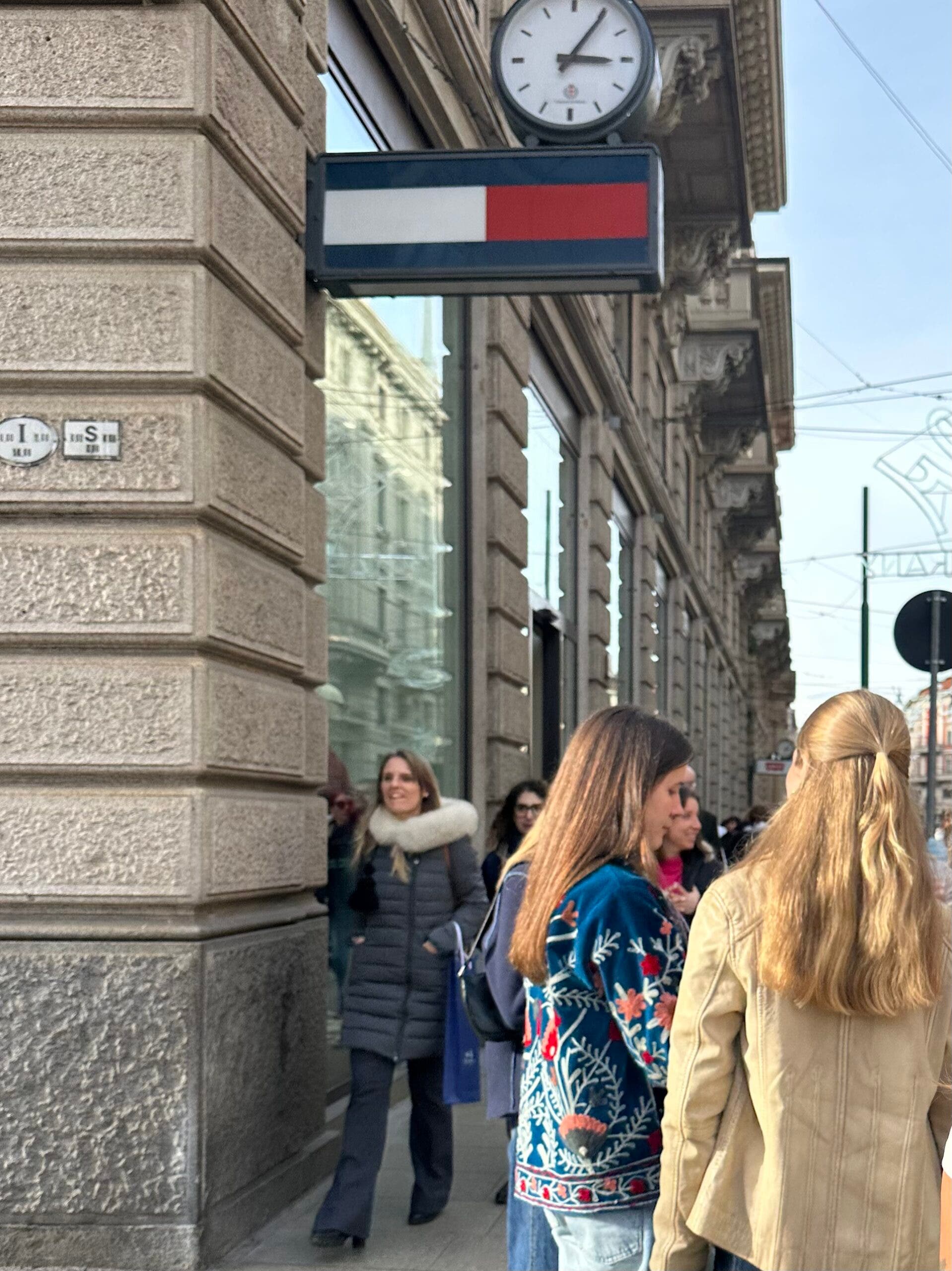 woman walks past a Tommy Hilfiger store in Milan wearing a vibrant blue jacket with floral embroidery. Another woman in a tan leather jacket stands in the foreground during the Milan-Cortina Winter Olympics.