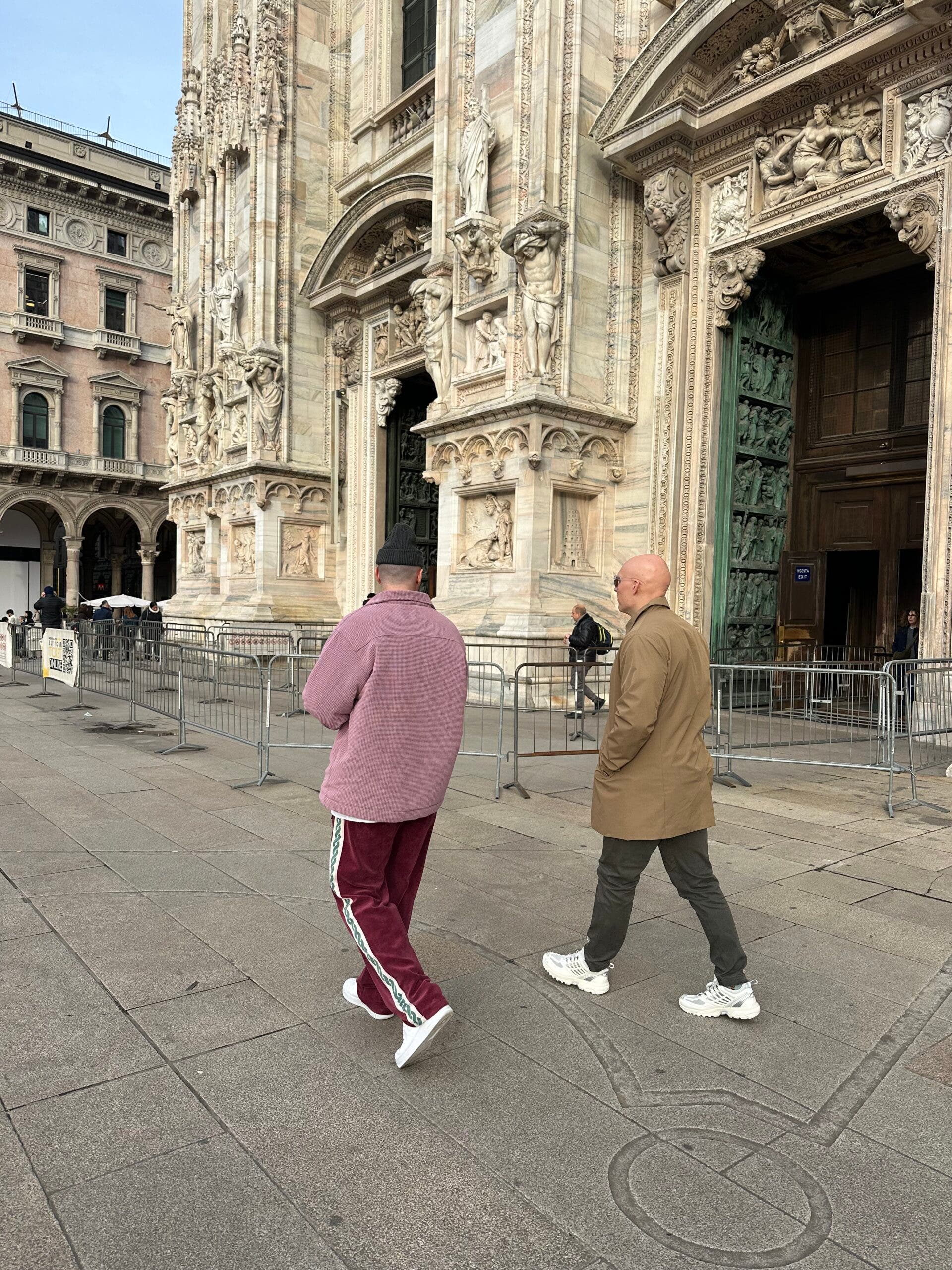 Two men walk past the intricate stone carvings of the Milan Cathedral (Duomo) during the Milan-Cortina Winter Olympics. One wears a pink corduroy overshirt and maroon track pants, while the other wears a tan trench coat.