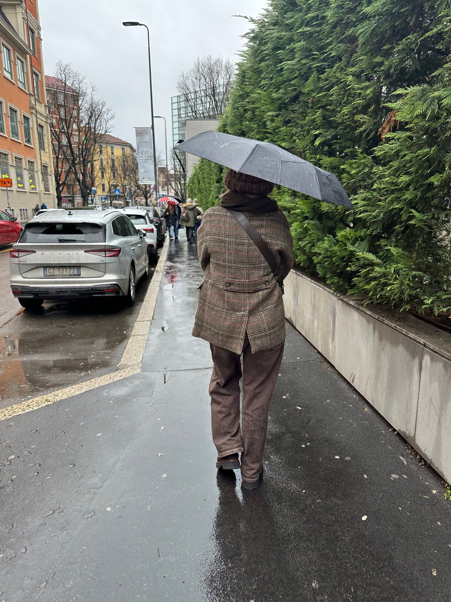 A person seen from behind walks down a rainy Milan street holding a black umbrella. They are wearing a brown plaid wool coat and brown trousers during the Milan-Cortina Winter Olympics.