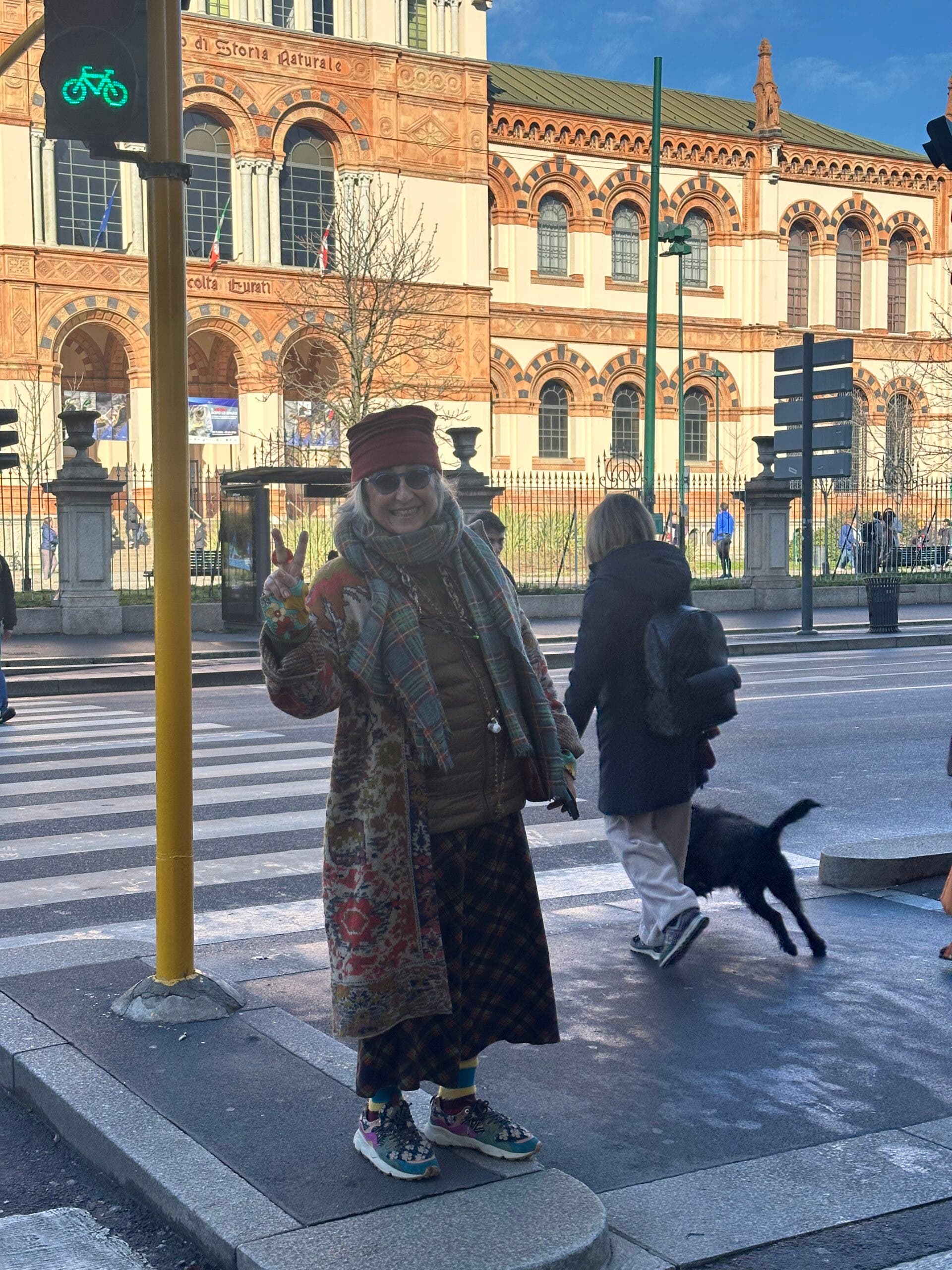 A smiling woman in Milan wears an eclectic mix of colorful patterns, a long tapestry-style coat, a plaid scarf, and a puffer vest. She gives a peace sign during the Milan-Cortina Winter Olympics.