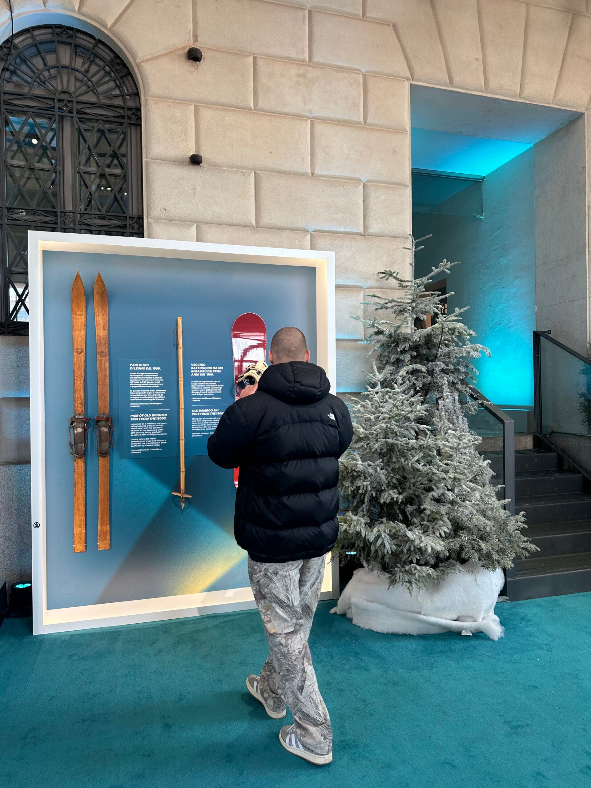 A man in a black North Face puffer jacket and camo pants looks at a museum display of vintage wooden skis and a red snowboard during the Milan-Cortina Winter Olympics.