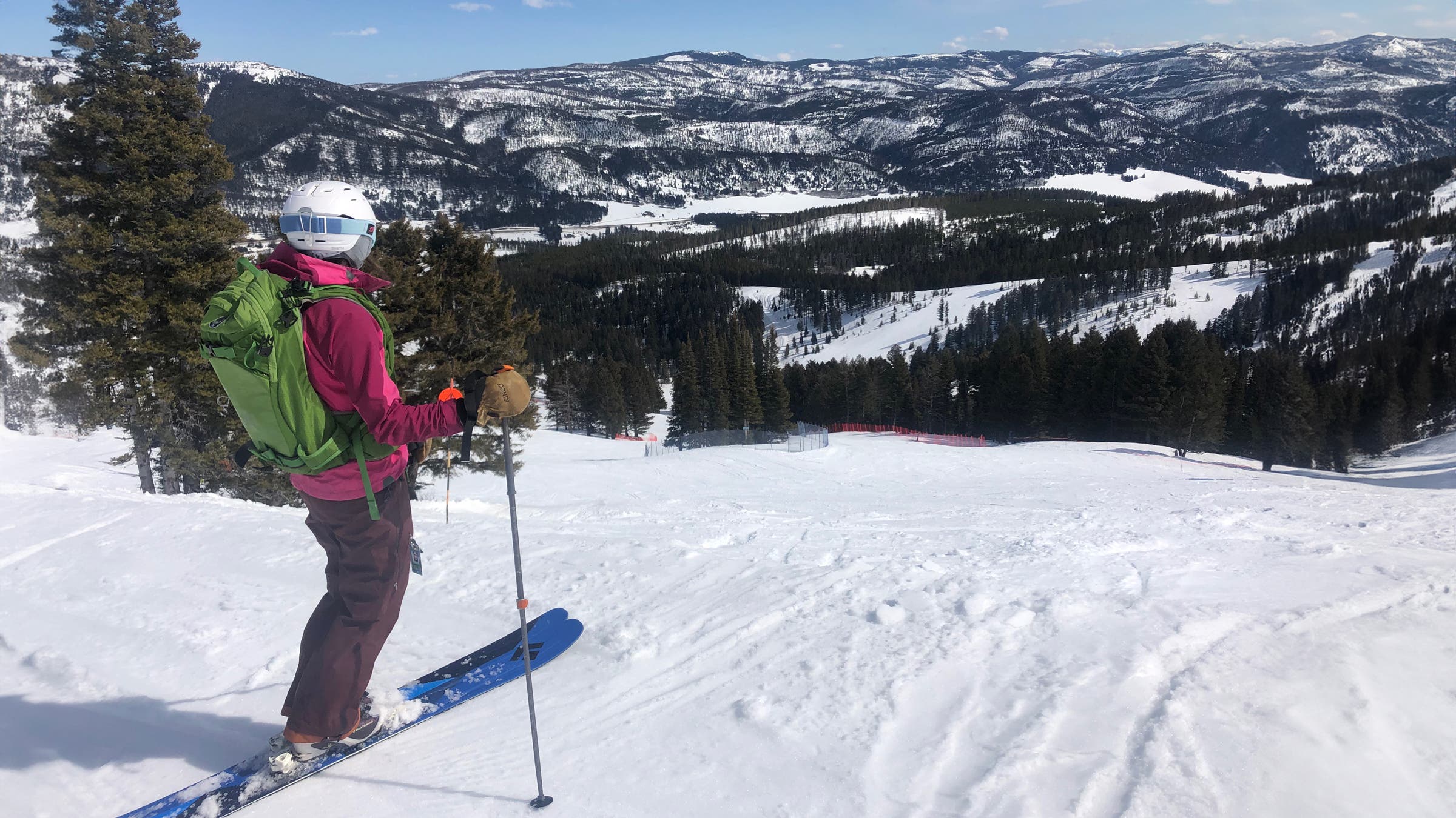 Skier in a pink jacket and green backpack pauses at the top of a snowy mountain run, wearing Kinco Grain & Suede leather mittens while looking out over forested slopes and distant snow-covered peaks.