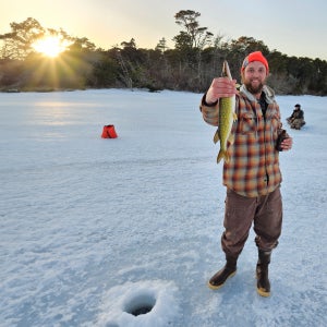 An angler stands on a frozen lake at sunset holding up a freshly caught fish beside an ice hole, wearing cold-weather layers and boots.