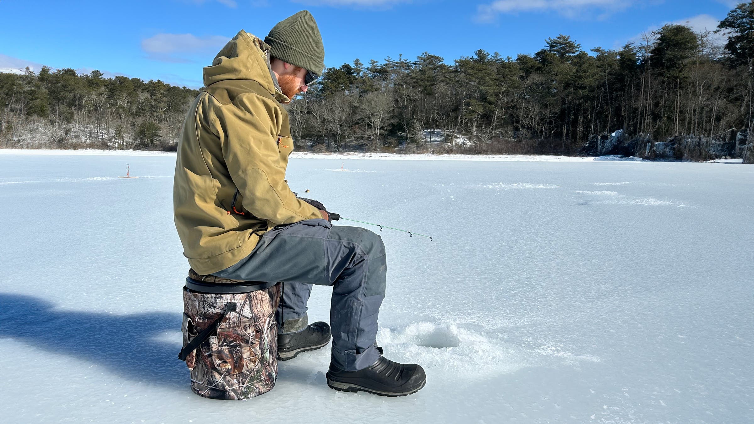 An angler sits on the WarBull 5 Gallon Bucket Swivel Seat while jigging over an ice hole on a frozen lake, wearing insulated boots and winter layers, highlighting practical ice fishing gear for comfort and mobility.