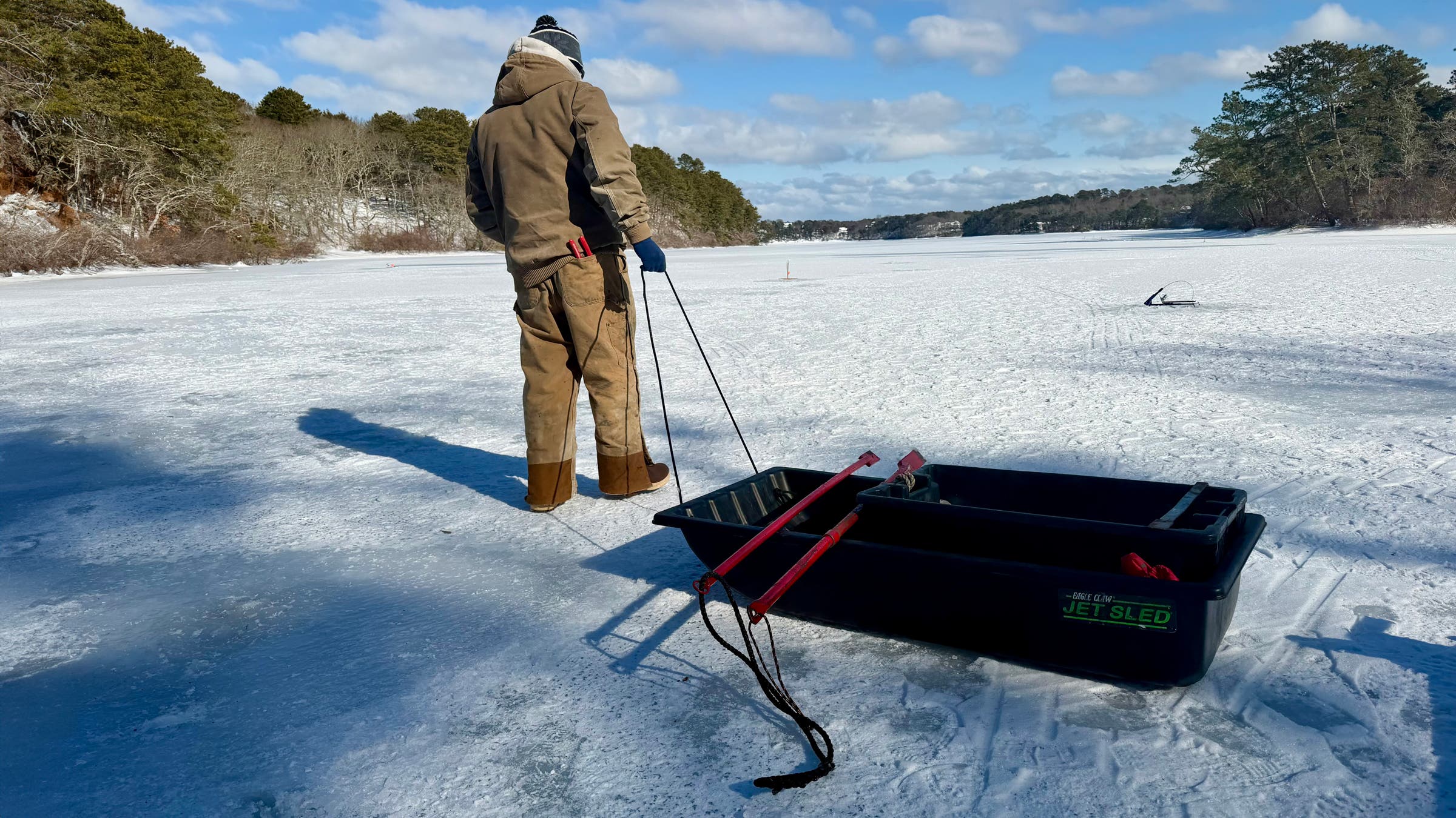An angler pulls a loaded Pelican Multi-Purpose Utility Sled across a frozen lake, transporting rods, tools, and other essential ice fishing gear over smooth ice under a bright winter sky.