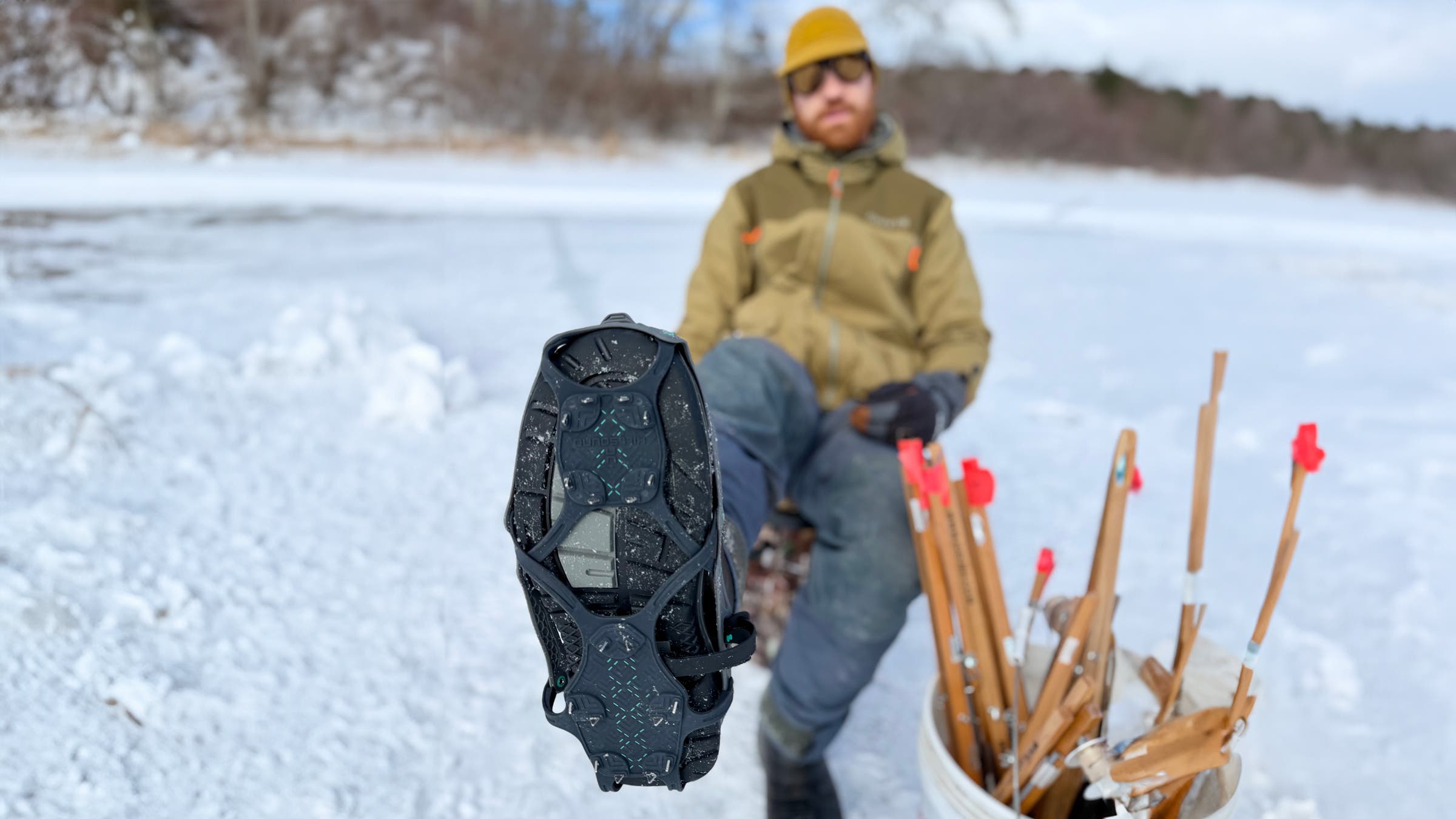 A close-up of the Hillsound FreeSteps6 All-Purpose Traction System strapped to a winter boot, shown sole-forward on a frozen lake with an angler and tip-ups in the background, highlighting durable ice fishing gear designed for grip and stability on slick ice.