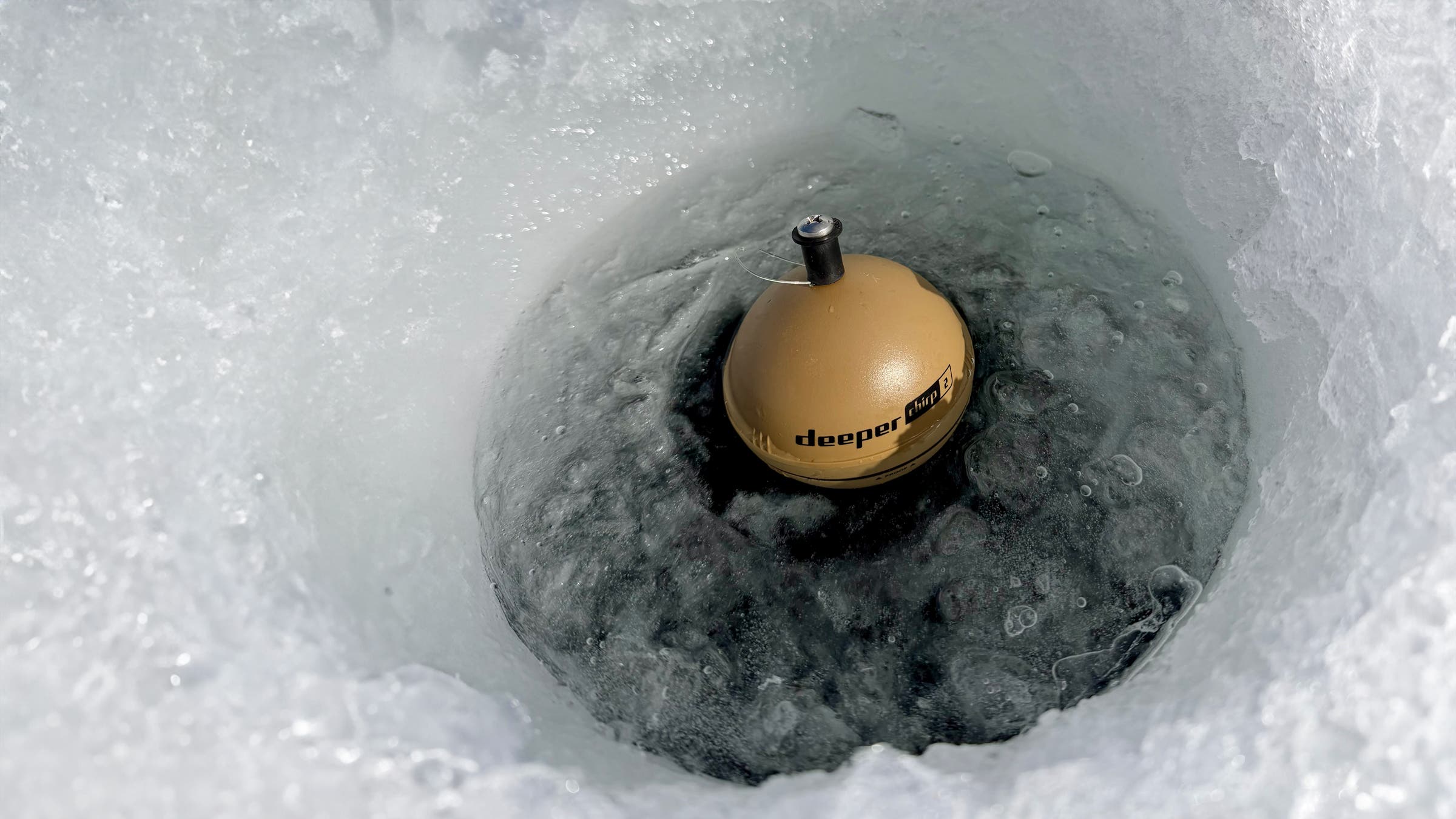A close-up view inside a freshly drilled ice hole shows the Deeper PRO+ 2 Portable Fish Finder floating in dark water, illustrating compact, high-tech ice fishing gear used to locate fish beneath frozen lakes.