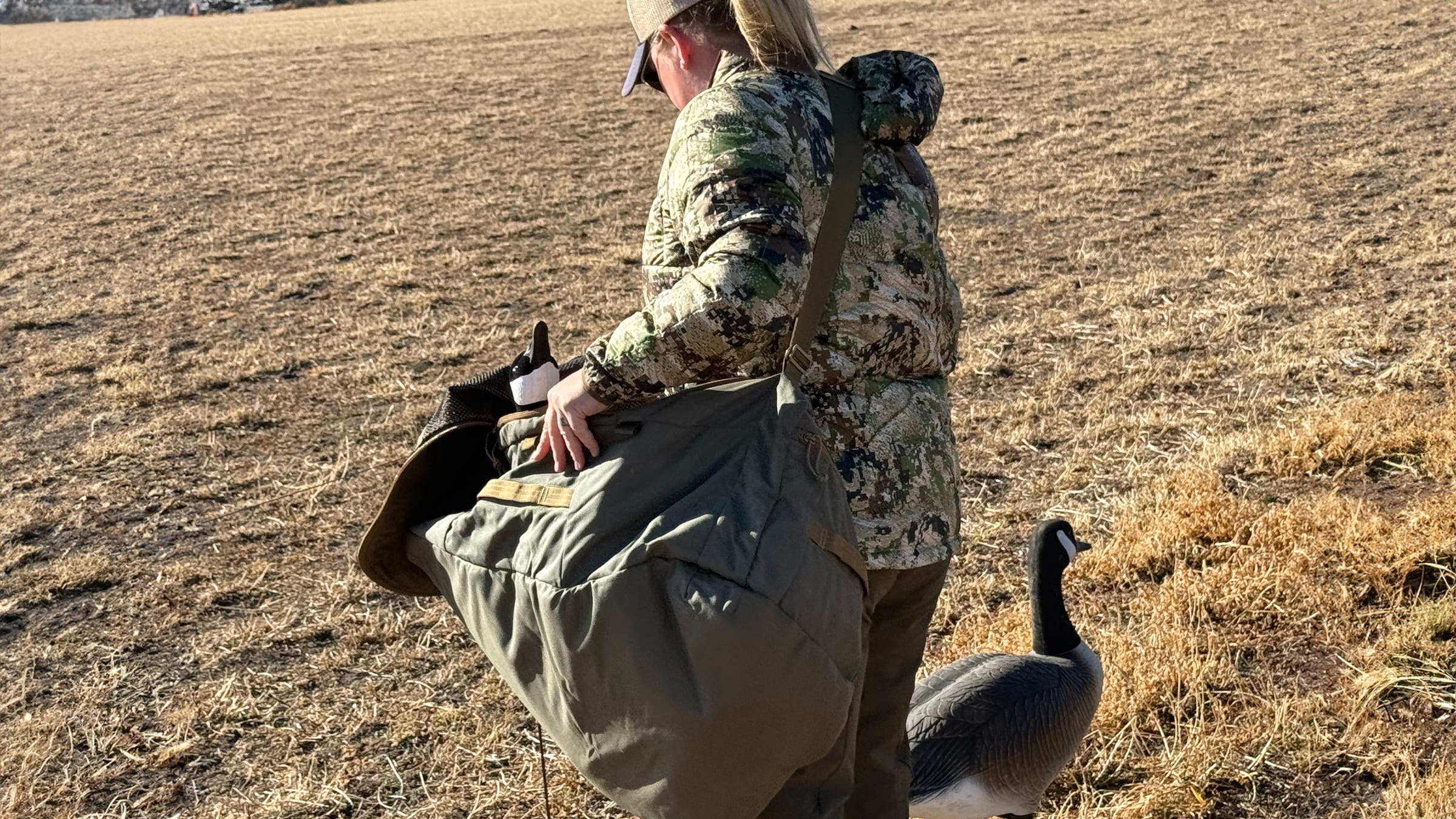 Man in camouflage jacket and trucker hat carrying a Kifaru Rondy Duffel in field.