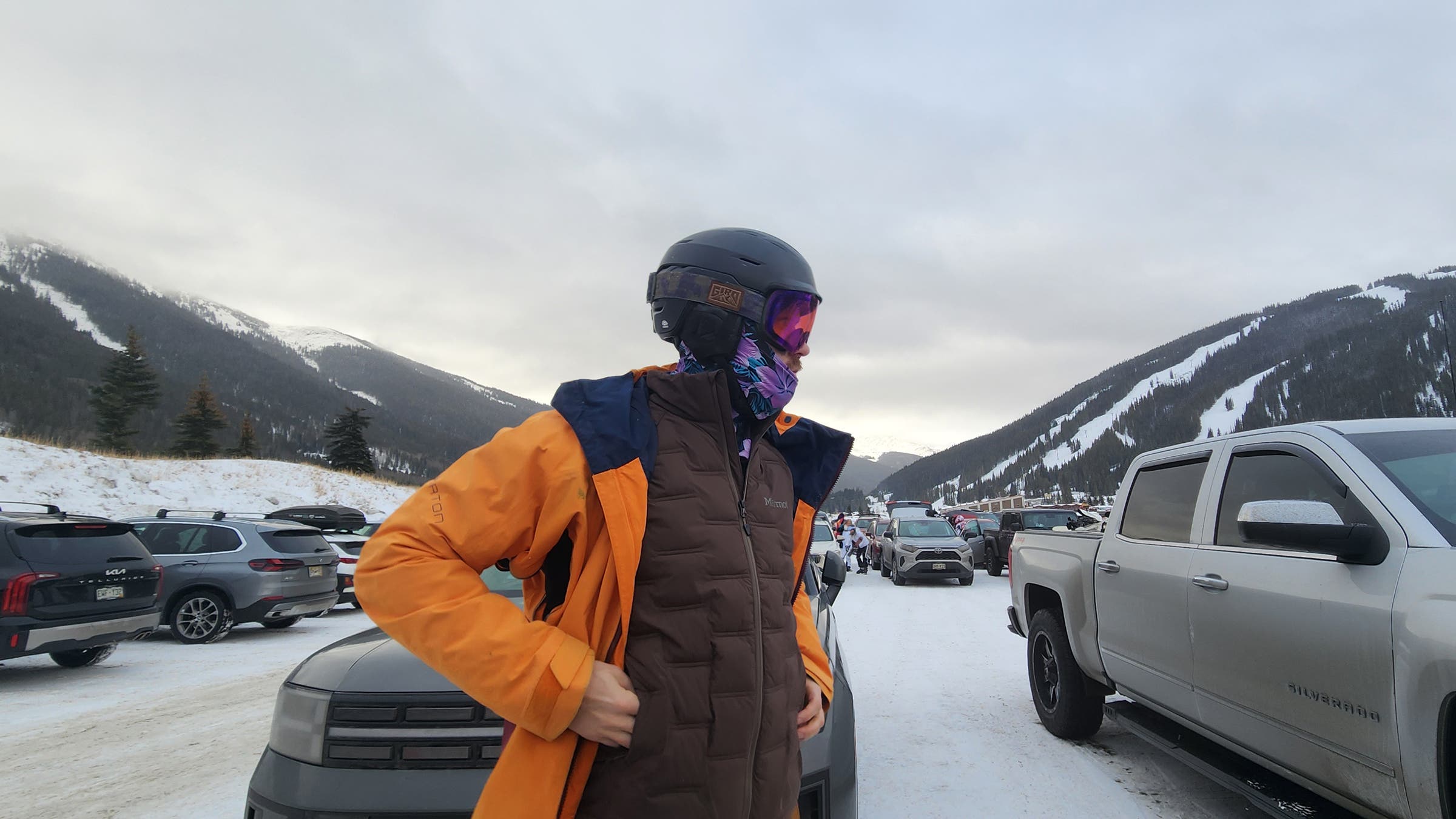 A skier adjusts layers in a snowy mountain parking lot while wearing the Marmot WarmCube Active Novus Insulated Jacket, a breathable synthetic jacket designed for active cold-weather use and efficient warmth on winter days.