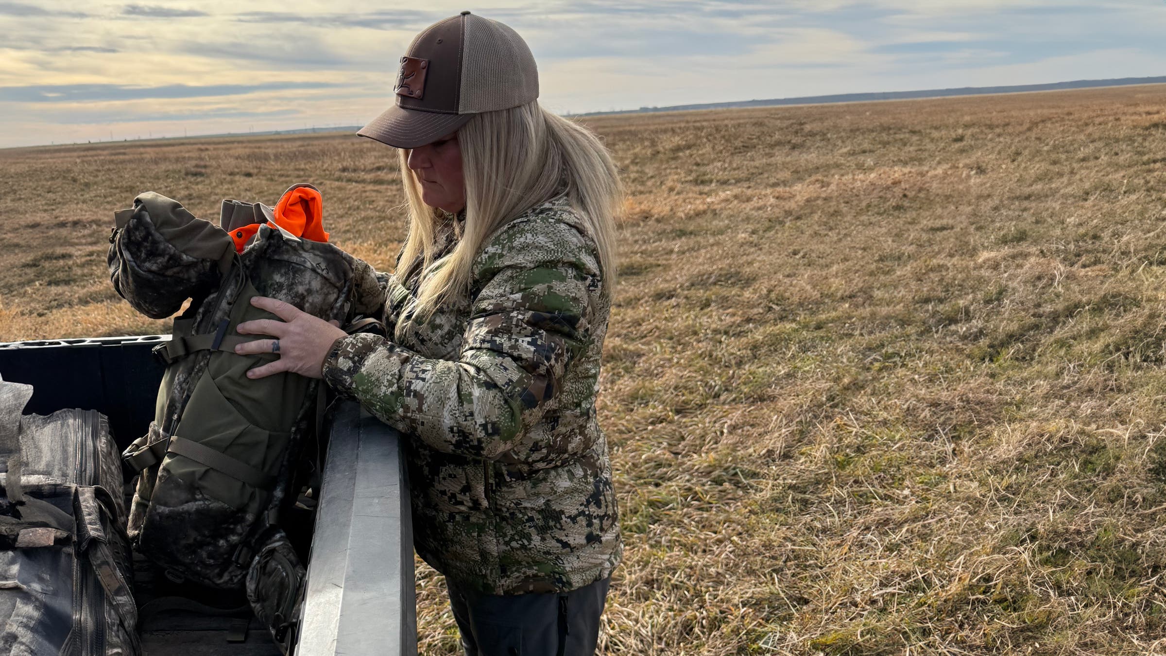 Woman in camouflage jacket and trucker hat loading a Kifaru Rondy Duffel into the bed of a pickup truck in an open prairie landscape, organizing hunting accessories and gear for a day in the field.