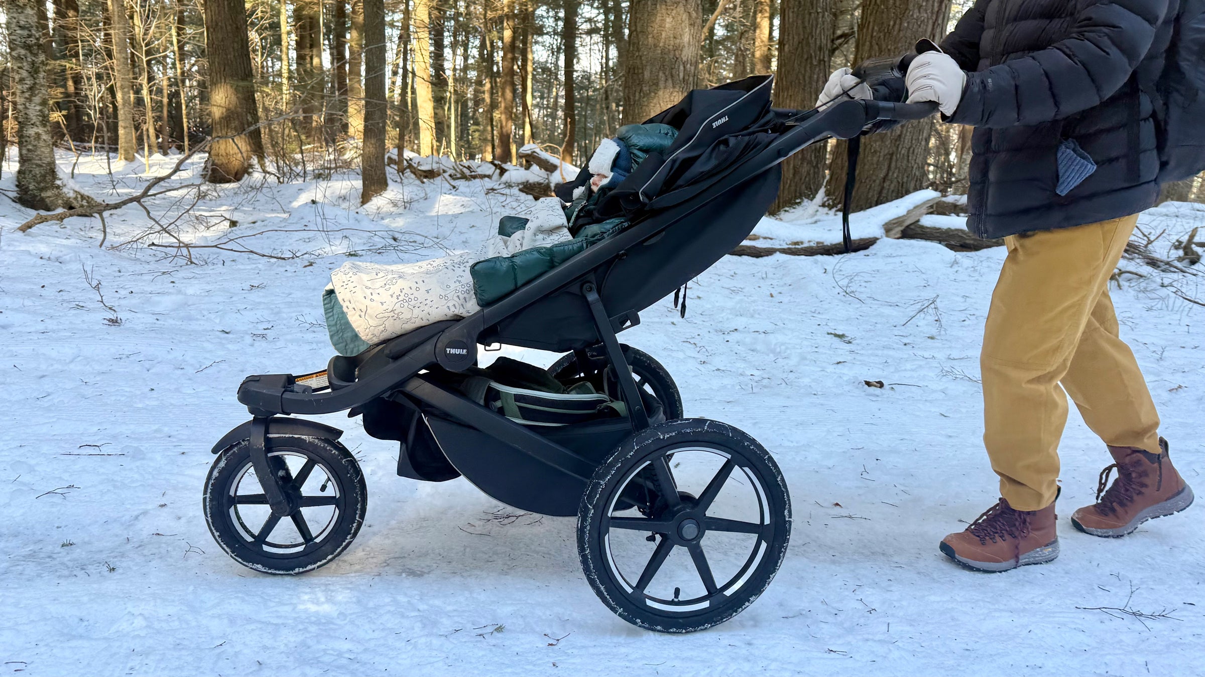 A parent pushes the Thule Urban Glide 3 jogging stroller along a snowy forest trail, showcasing its large air-filled tires and sturdy three-wheel design as a bundled baby rides reclined and warm beneath the canopy.