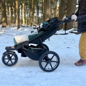 A parent pushes the Thule Urban Glide 3 jogging stroller along a snowy forest trail, showcasing its large air-filled tires and sturdy three-wheel design as a bundled baby rides reclined and warm beneath the canopy.