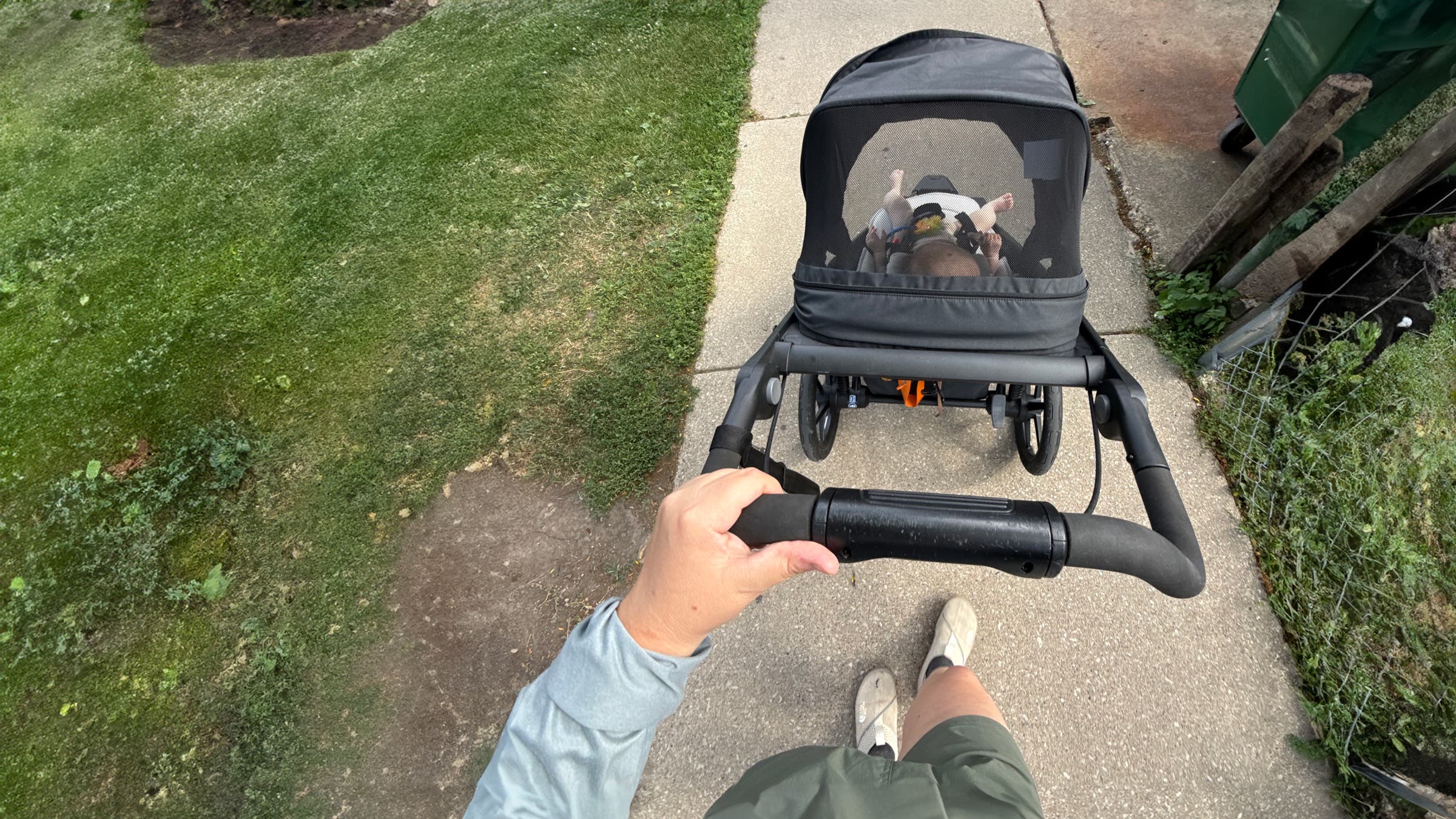 First-person view of a parent pushing the Thule Urban Glide 3 jogging stroller along a neighborhood sidewalk, showing the ergonomic handlebar, mesh canopy window, and three-wheel design as a baby rides comfortably inside.