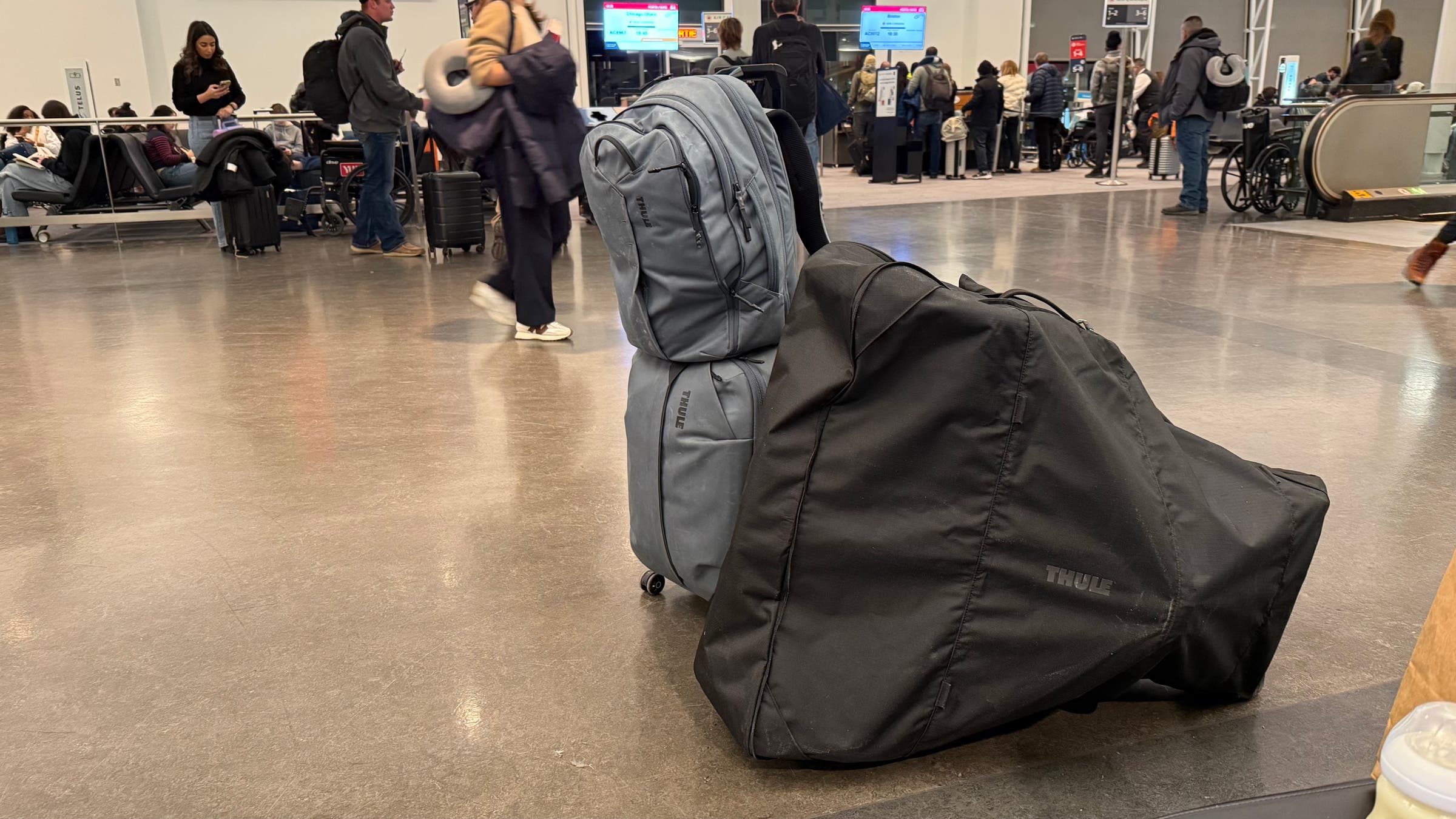 A packed Thule Urban Glide 3 jogging stroller sits inside a black travel bag at an airport terminal, leaning against rolling luggage and a backpack near a check-in counter, highlighting its portability for air travel.