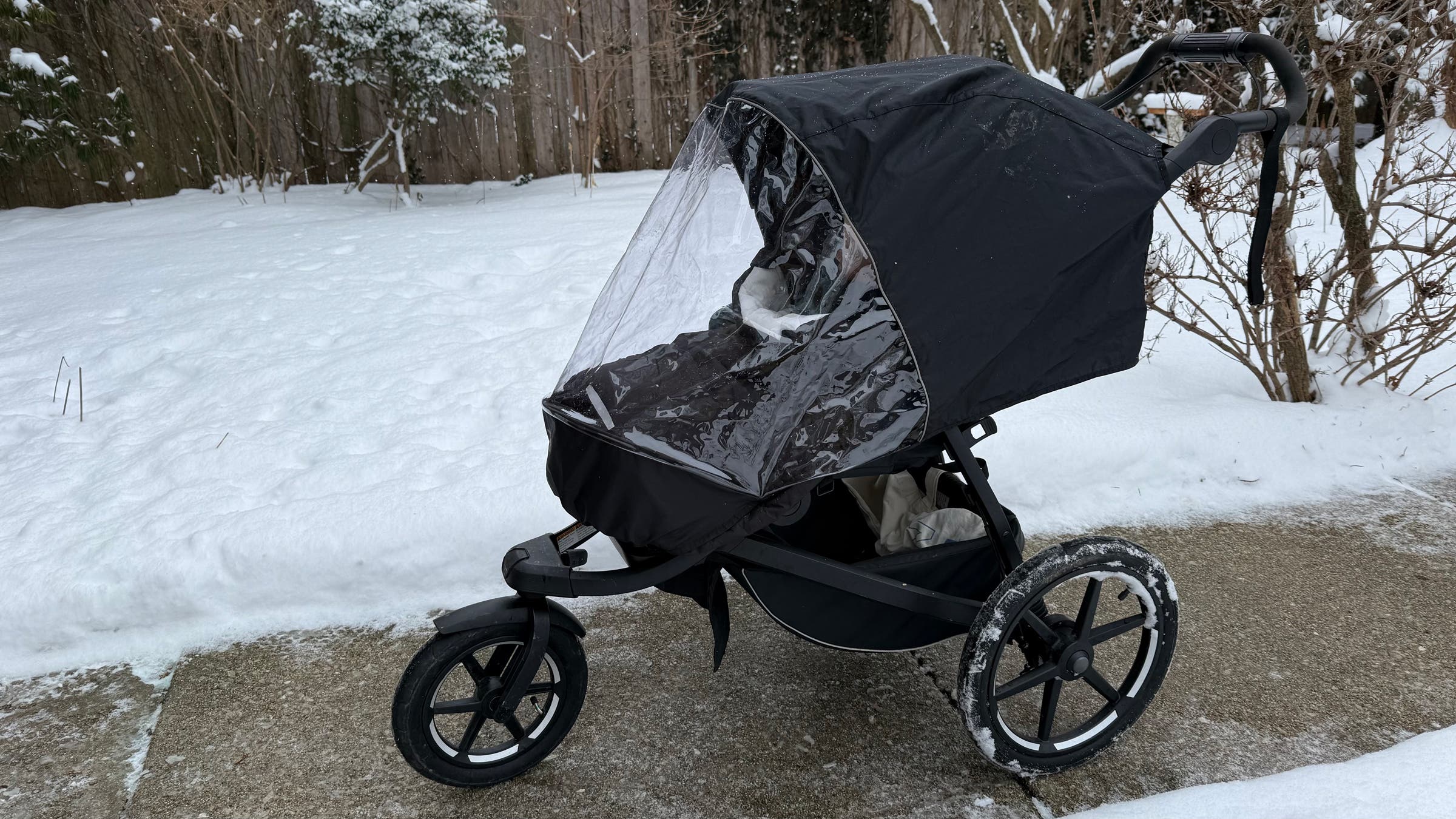 The Thule Urban Glide 3 jogging stroller sits on a cleared sidewalk beside snowbanks, fitted with a transparent rain cover over the canopy to shield a bundled baby, highlighting its three-wheel design and winter-ready versatility.