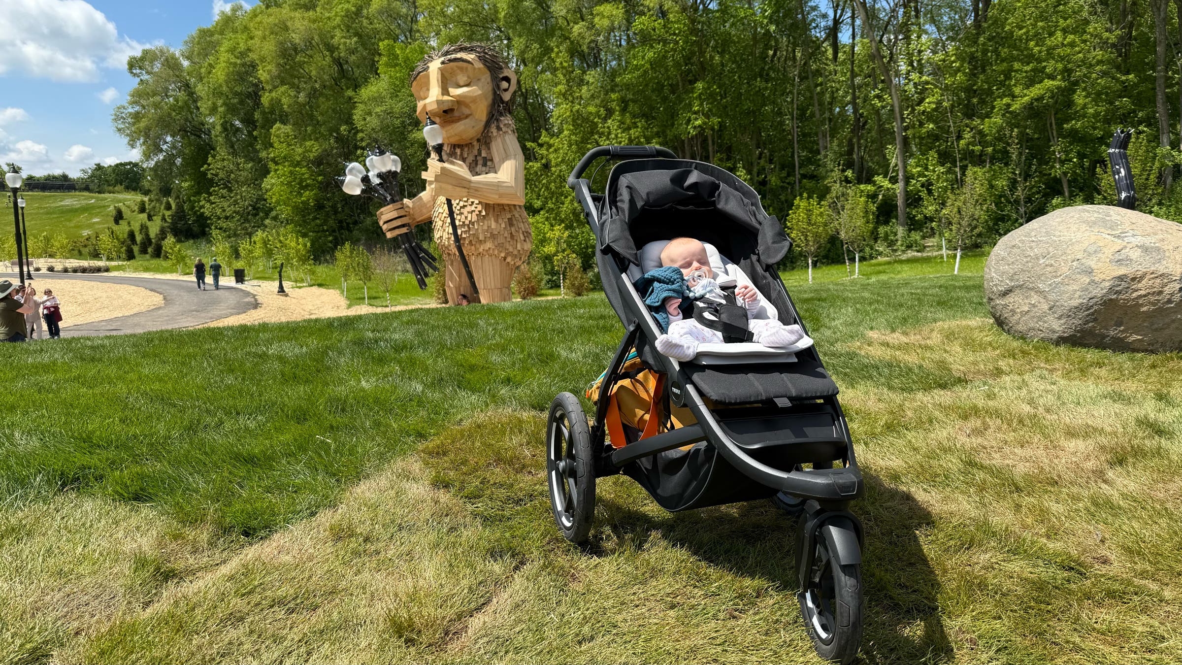 A baby naps in the Thule Urban Glide 3 jogging stroller parked on a grassy park lawn, with a large wooden troll sculpture and tree-lined path in the background, highlighting the stroller’s three-wheel design and all-terrain capability for outdoor adventures.