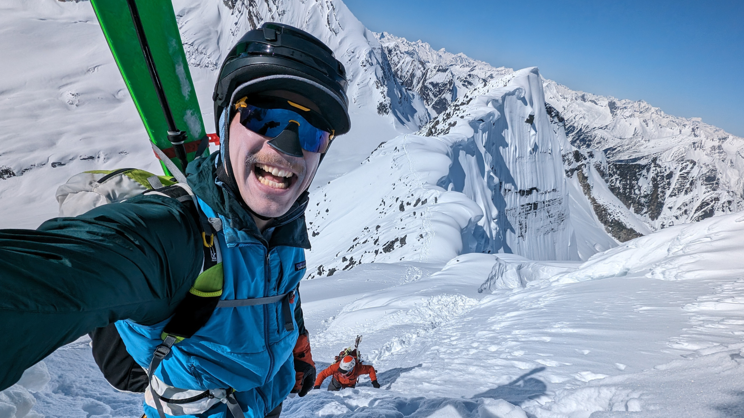 A smiling skier with a skimo pack and skis attached, taking a selfie while ascending a steep, snowy mountain.