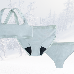 A light blue bralette and two pairs of period proof underwear atop a snowy forest background