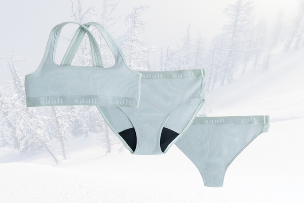 A light blue bralette and two pairs of period proof underwear atop a snowy forest background