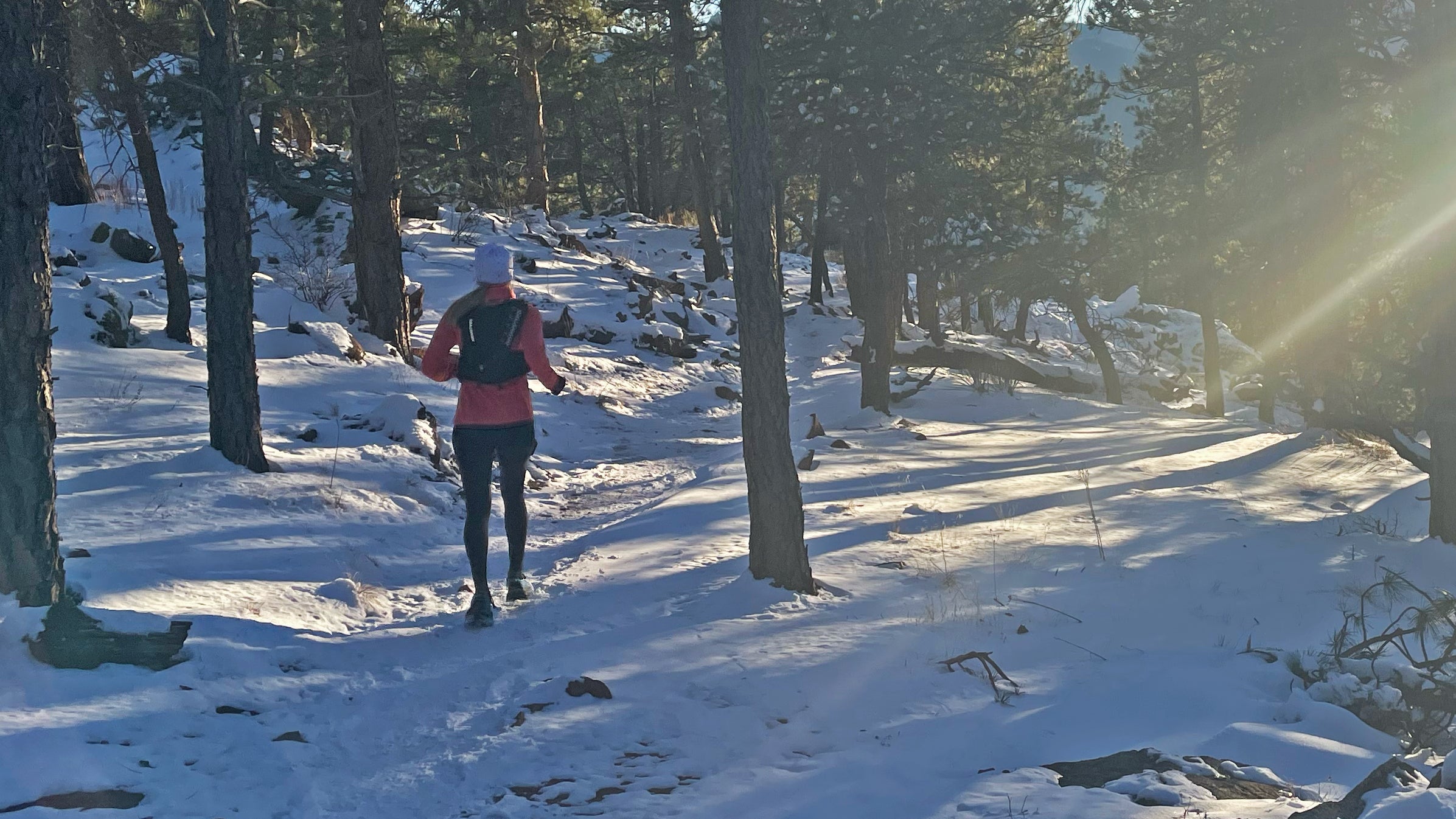 Author Lisa Jhung runs along a snowy forest trail wearing cold weather running gear, including a pink insulated layer, black tights, gloves, a beanie, and a hydration vest, as winter sunlight filters through tall pine trees.