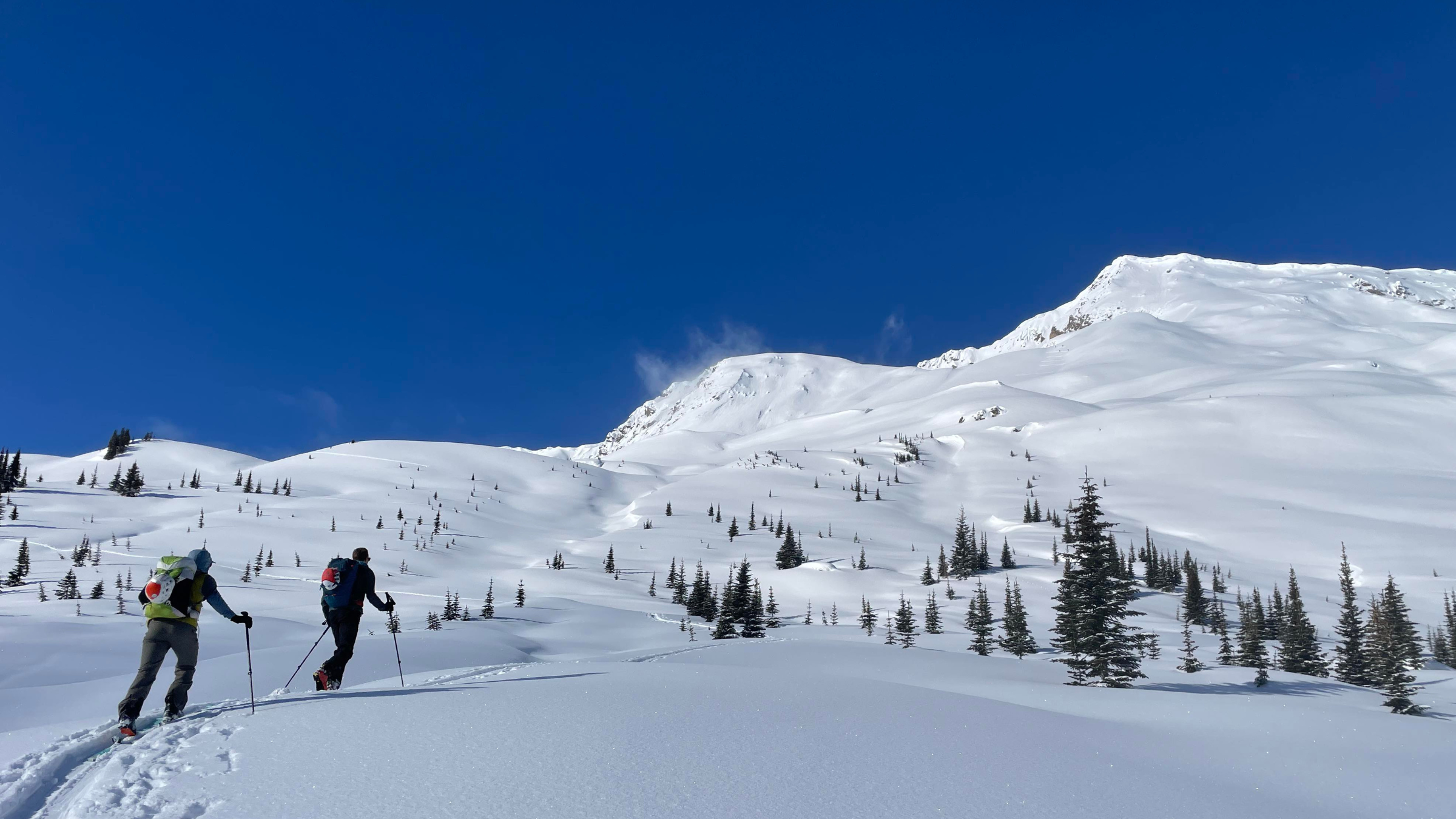 Two skiers using a lightweight skimo pack to climb a snowy mountain ridge under a clear blue sky.