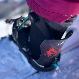 Close-up of a snowboard boot locked into the Jones Mercury FASE Snowboard Binding, showing the highback and dial-style FASE system set against a snowy mountain backdrop.