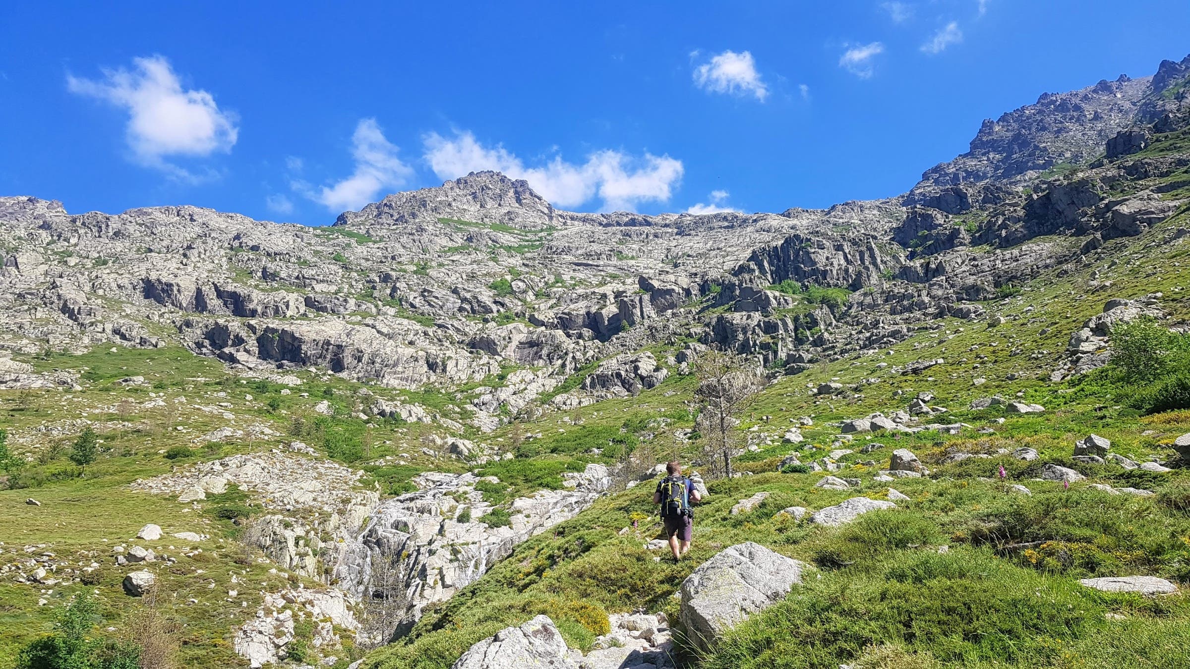 A lone hiker treks up a green and rocky mountainside.
