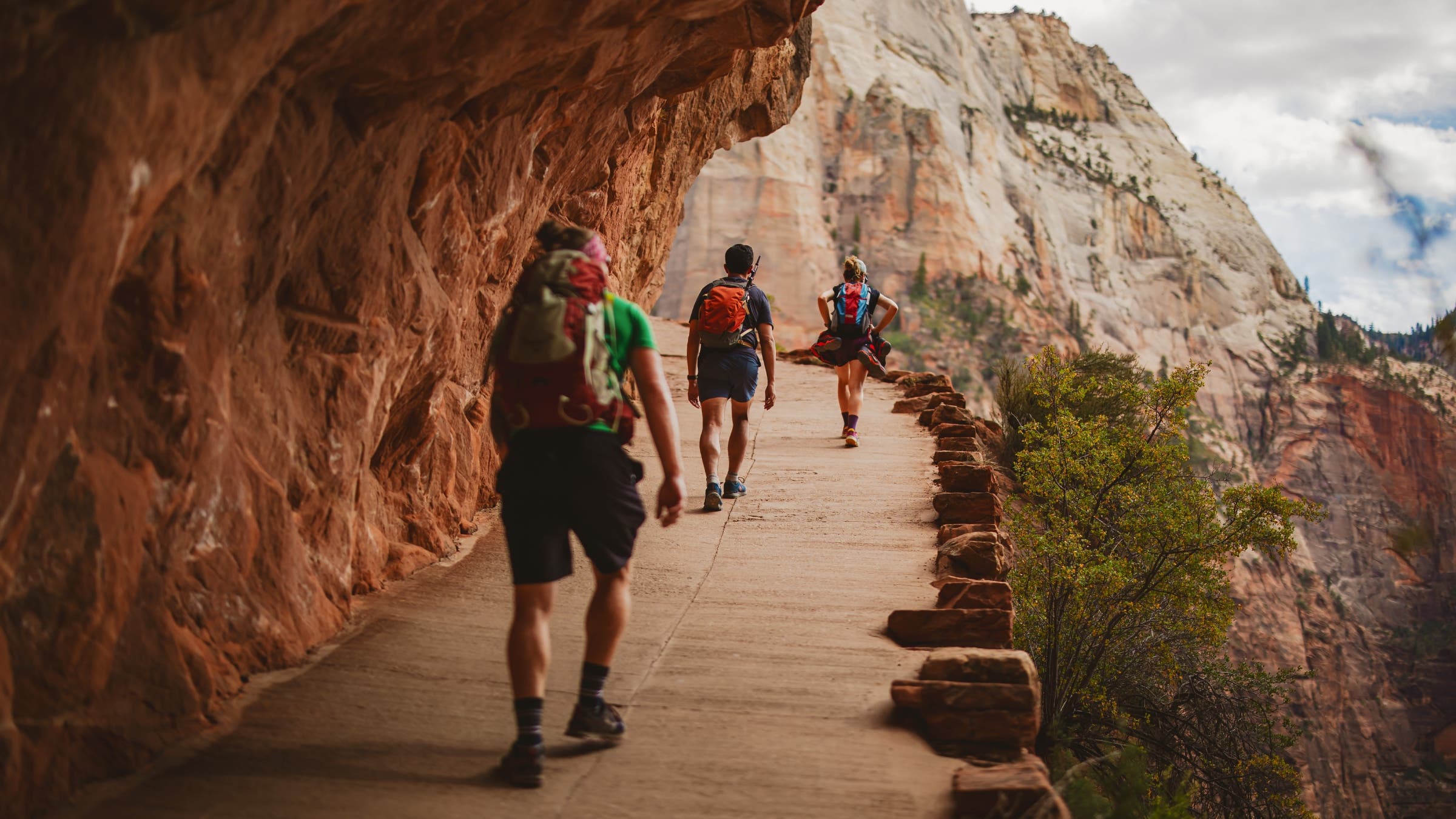 Hikers walk along a paved path carved into a red-rock cliff face.